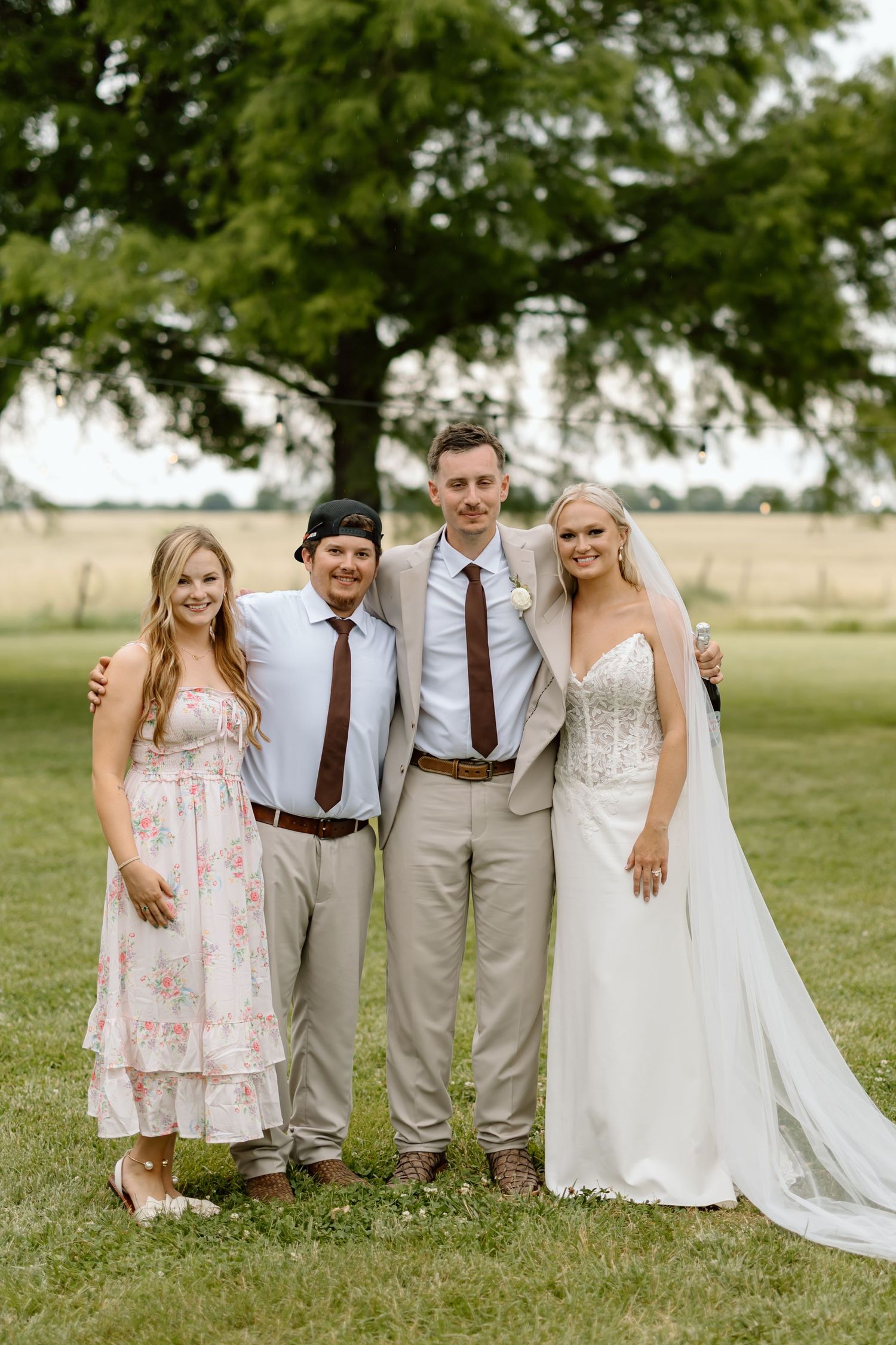 Wedding party of four posing outdoors on green grass, bride in white gown with veil, guests in formal attire near large tree.