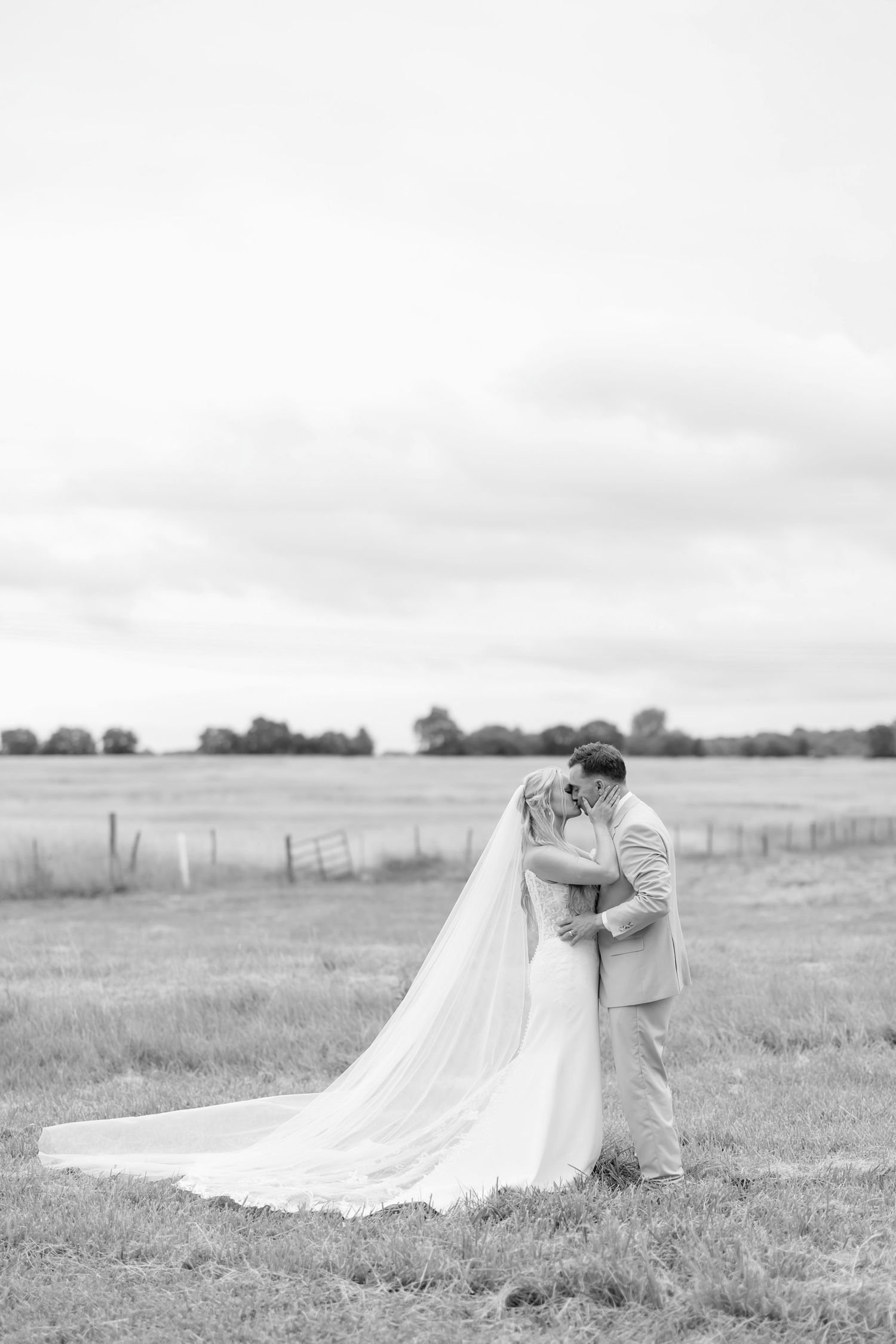 Black and white photo of a bride and groom sharing a romantic kiss in an open field on their wedding day.