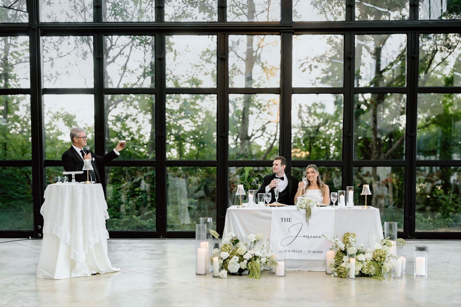 Elegant wedding reception with couple at sweetheart table, floral arrangements, candles, and large glass windows overlooking greenery.