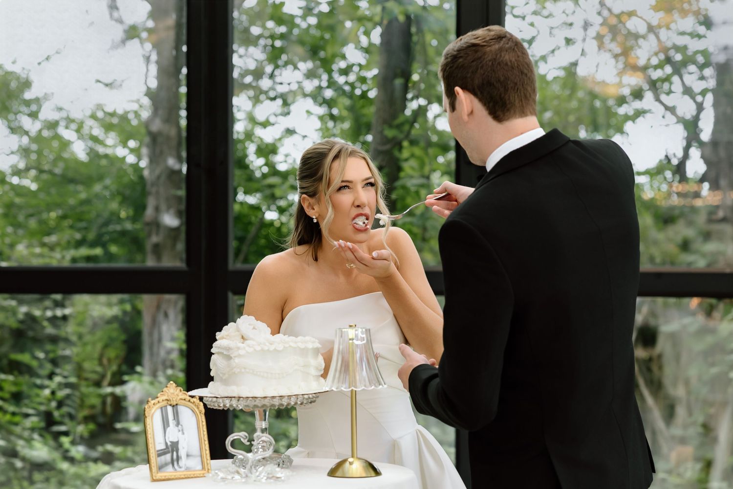 Bride and groom cutting wedding cake together indoors, bride smiling as groom feeds her cake at elegant reception.