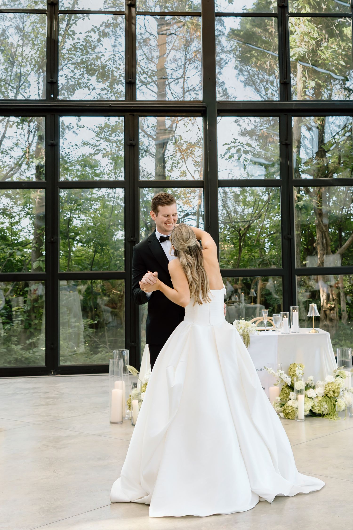 Bride and groom share their first dance in an elegant venue with floor-to-ceiling windows and lush greenery backdrop.