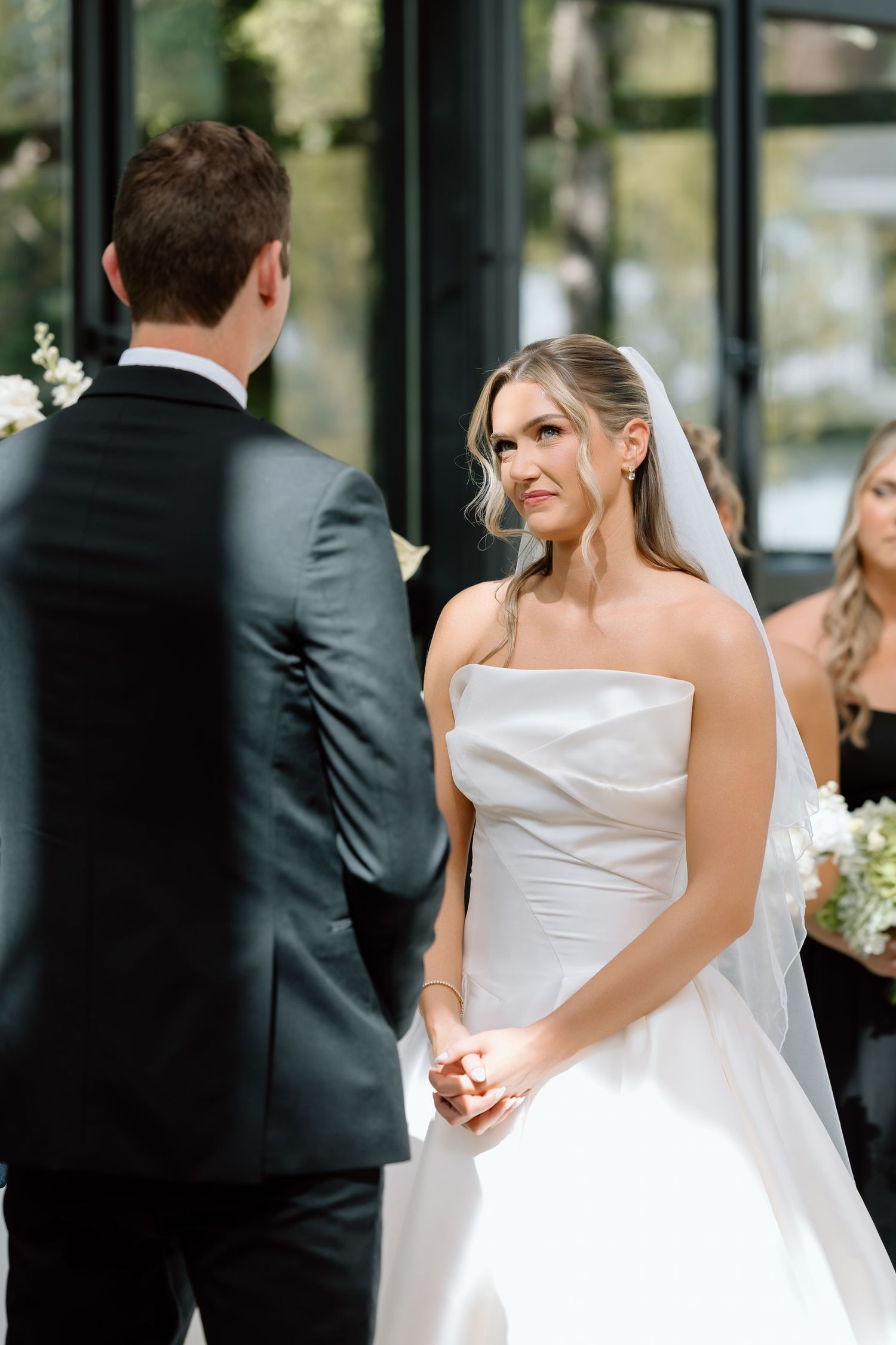 Bride in strapless white gown holds hands with groom in black tuxedo during outdoor wedding ceremony.