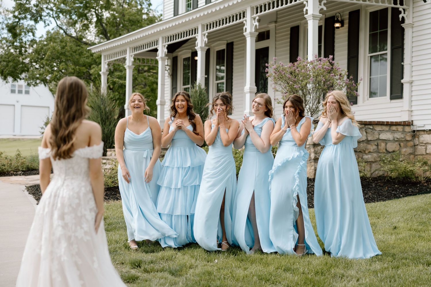Bridesmaids in light blue gowns react with joy seeing the bride in a white lace dress outside a white farmhouse.