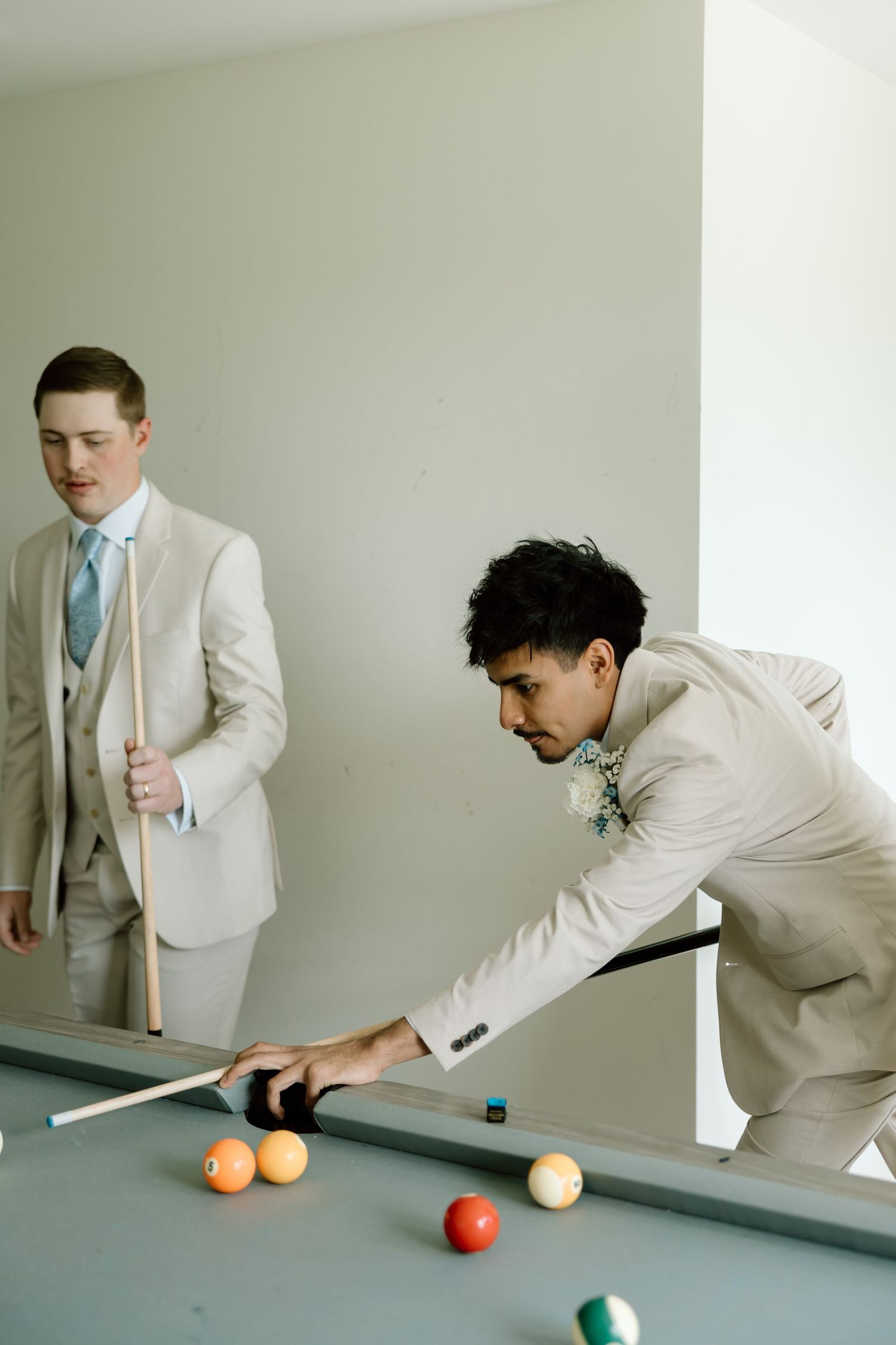 Two groomsmen in beige suits playing pool, one leaning over the billiard table to take a shot.