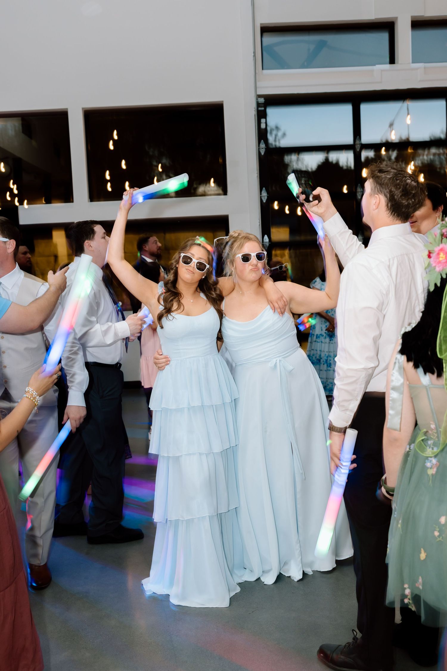 Two bridesmaids in light blue gowns and sunglasses celebrate with glowing foam sticks at a festive wedding reception.