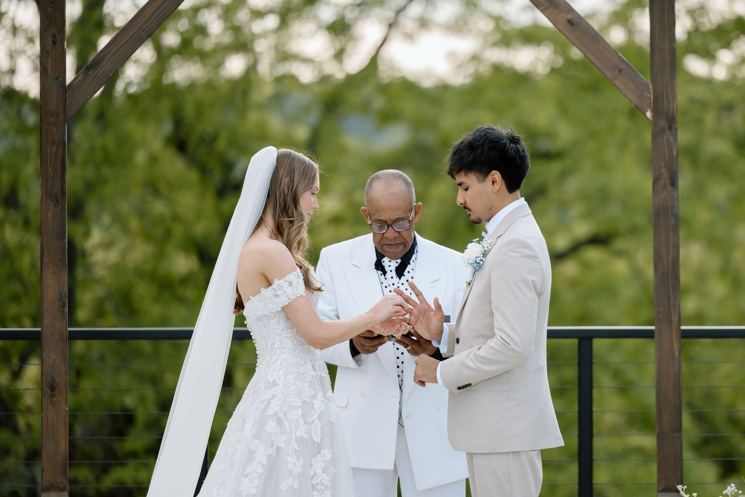 Bride and groom exchange rings during outdoor wedding ceremony with officiant under wooden arch.