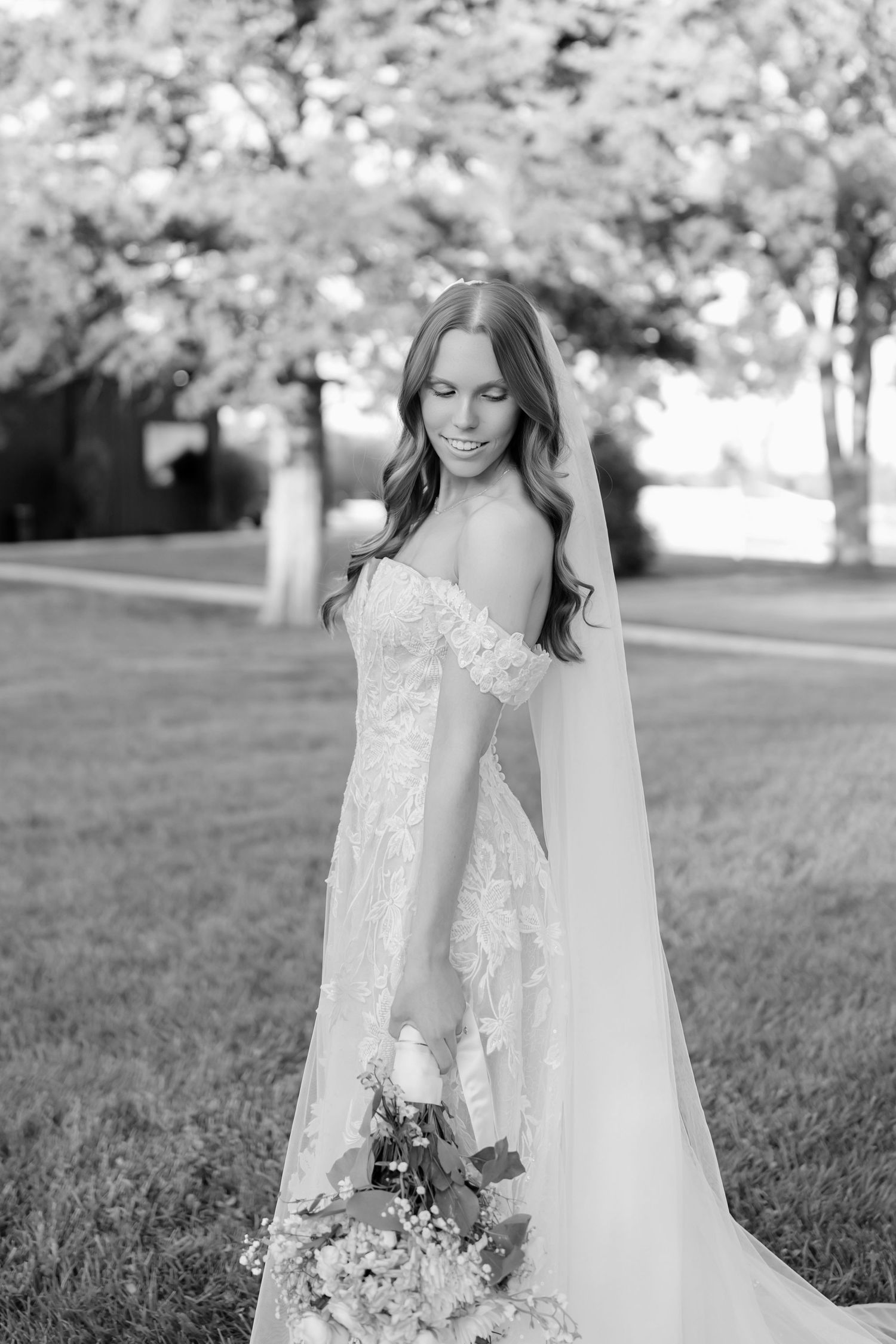 Black and white photo of a bride in lace off-shoulder gown holding bouquet, standing outdoors on grass with trees.