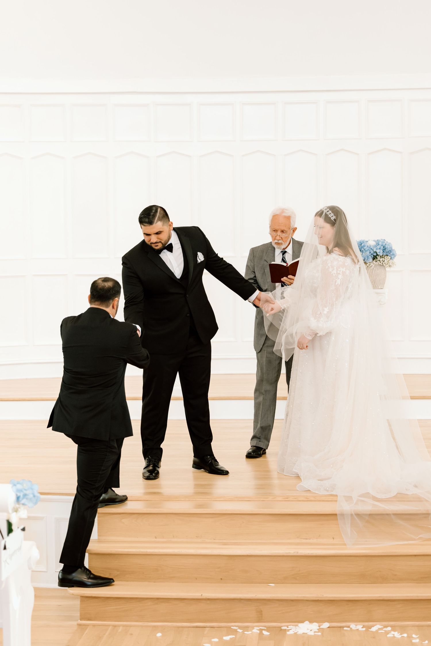 Groom in black tuxedo holds bride's hand at wedding altar with officiant holding book in white chapel.