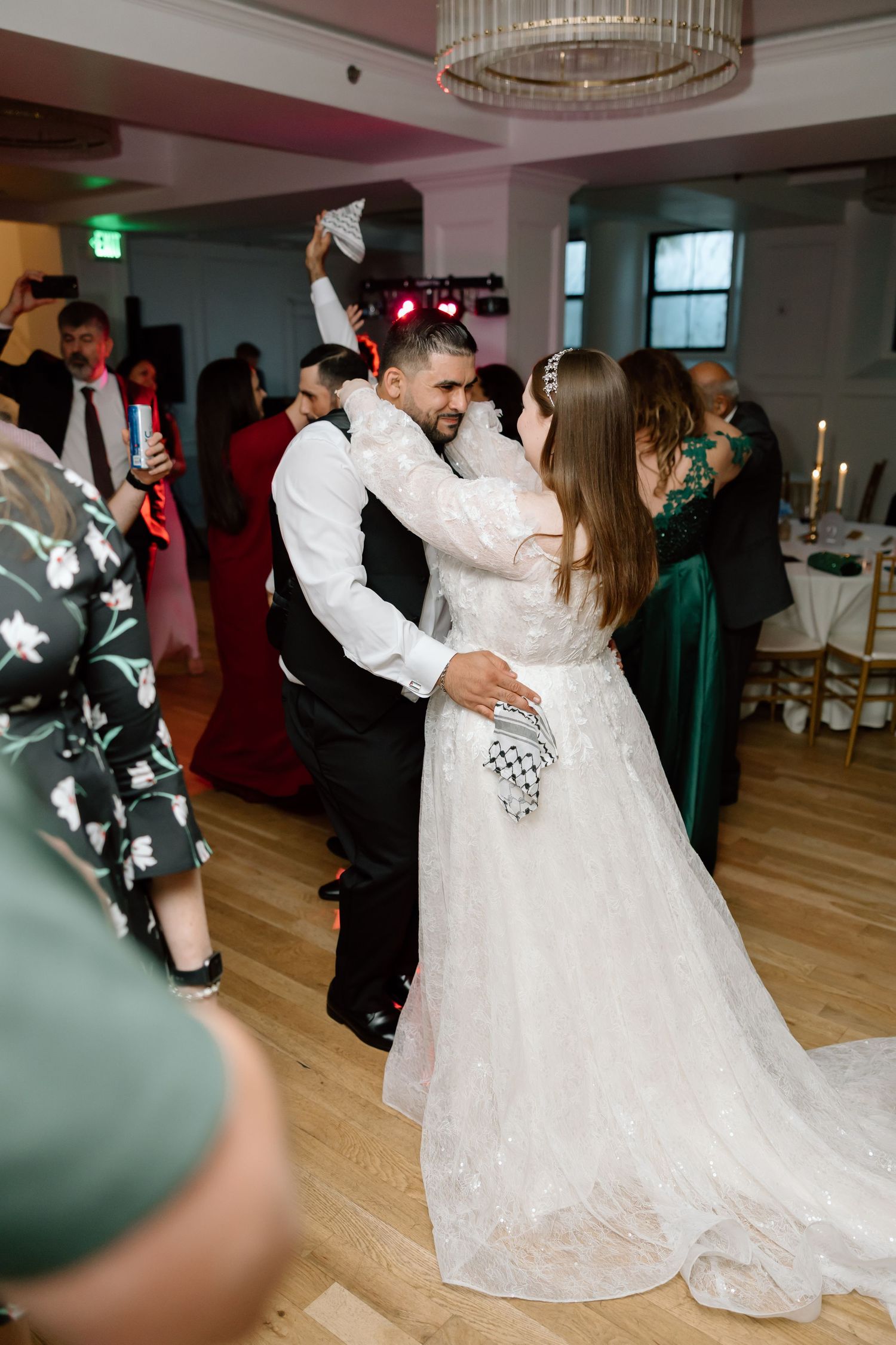 Bride and groom share first dance at wedding reception, surrounded by guests in elegant venue with chandelier lighting.