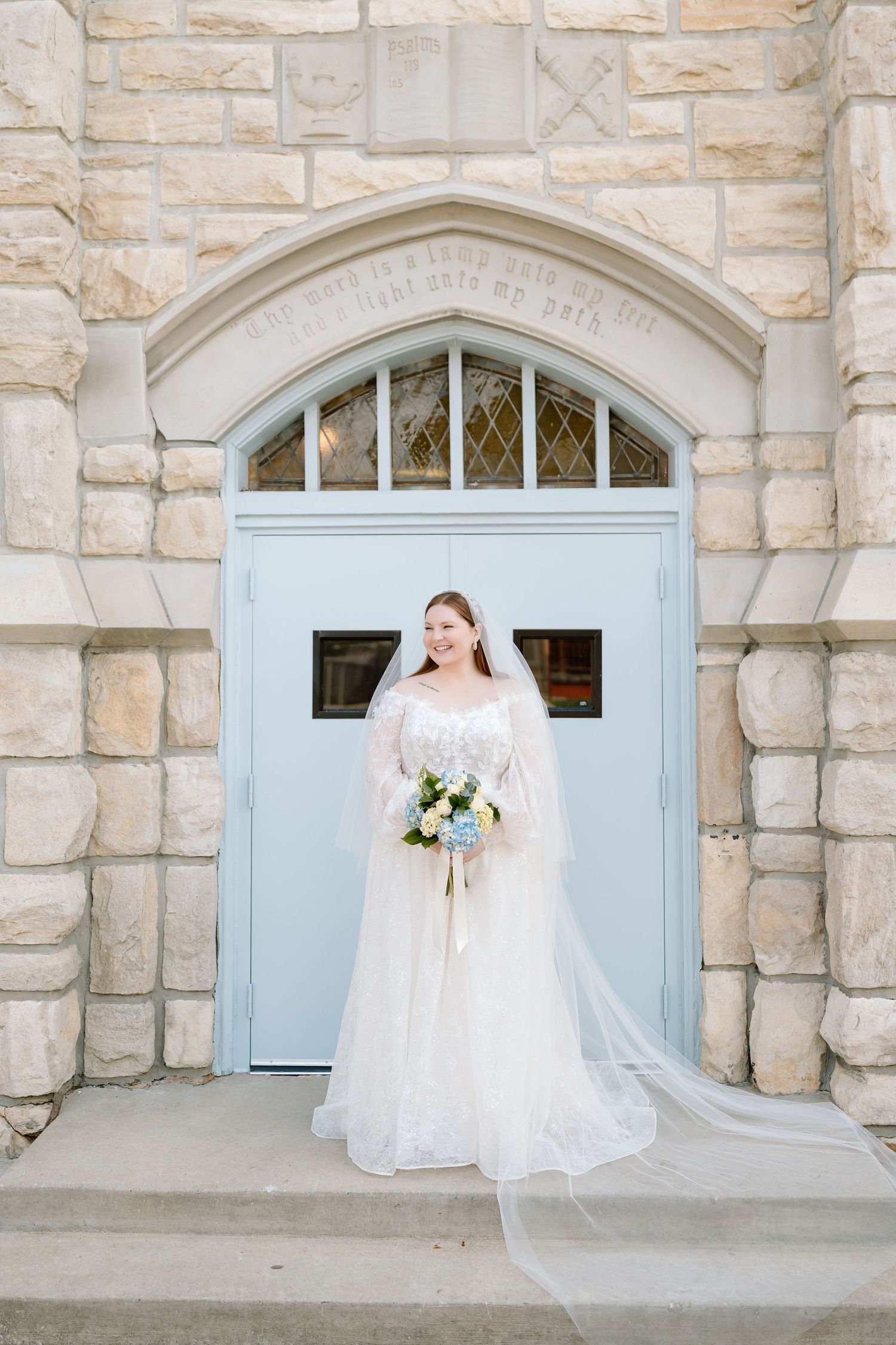Bride in lace gown and veil holds bouquet before stone church's blue arched door.