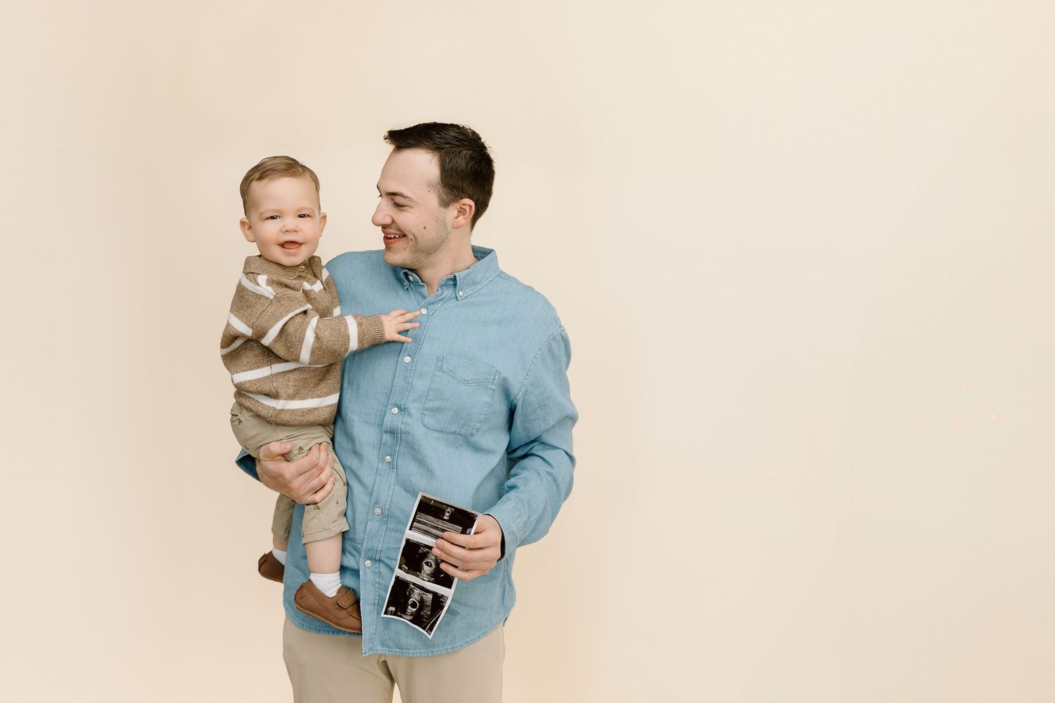 A parent in blue holds a small child wearing brown while looking at photos together against a light background.