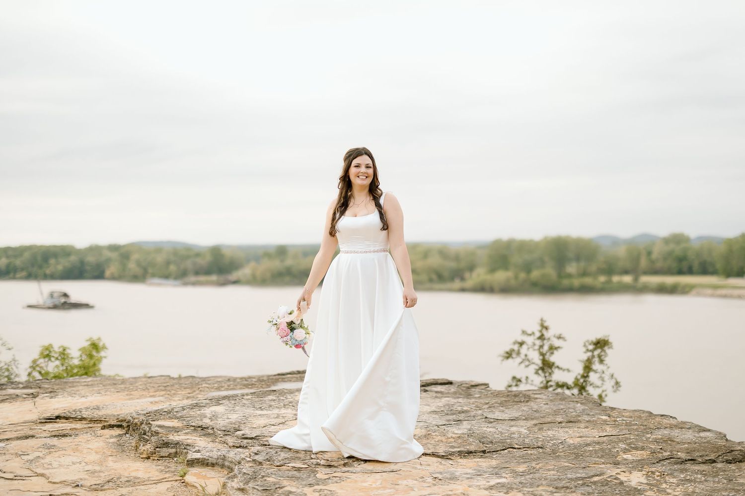 A bride in a flowing white dress stands on a rocky overlook with a serene lake and trees in the background.
