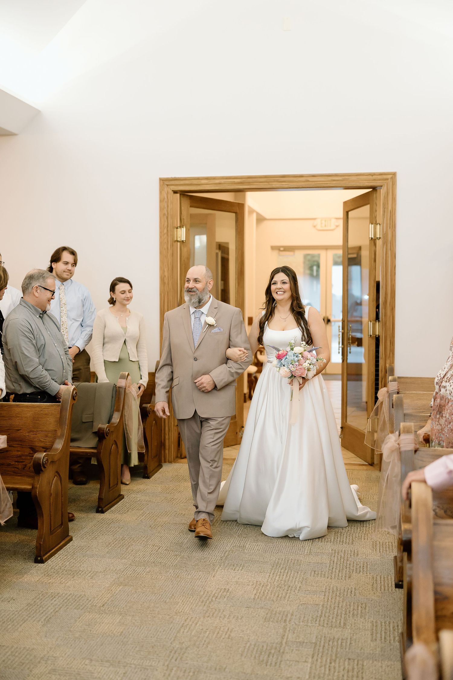 A couple walks down the aisle of a chapel during a wedding ceremony with wooden pews and guests looking on.