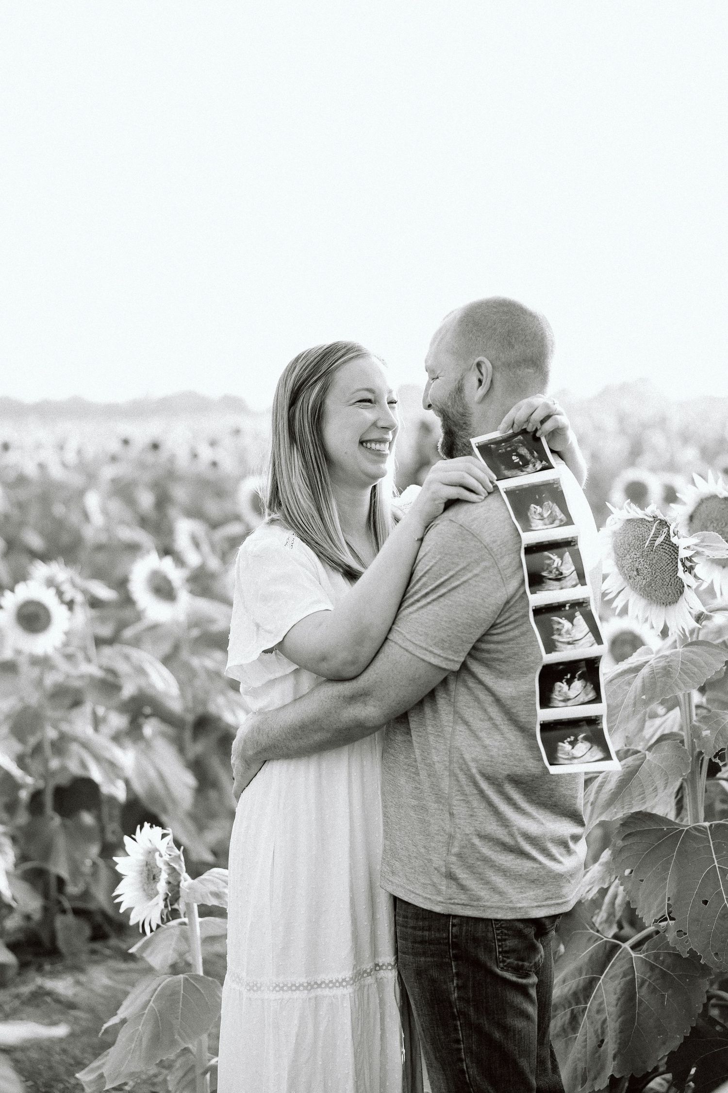 A couple in a sunflower field shares a joyful moment while looking at ultrasound photos in a series of black and white images.