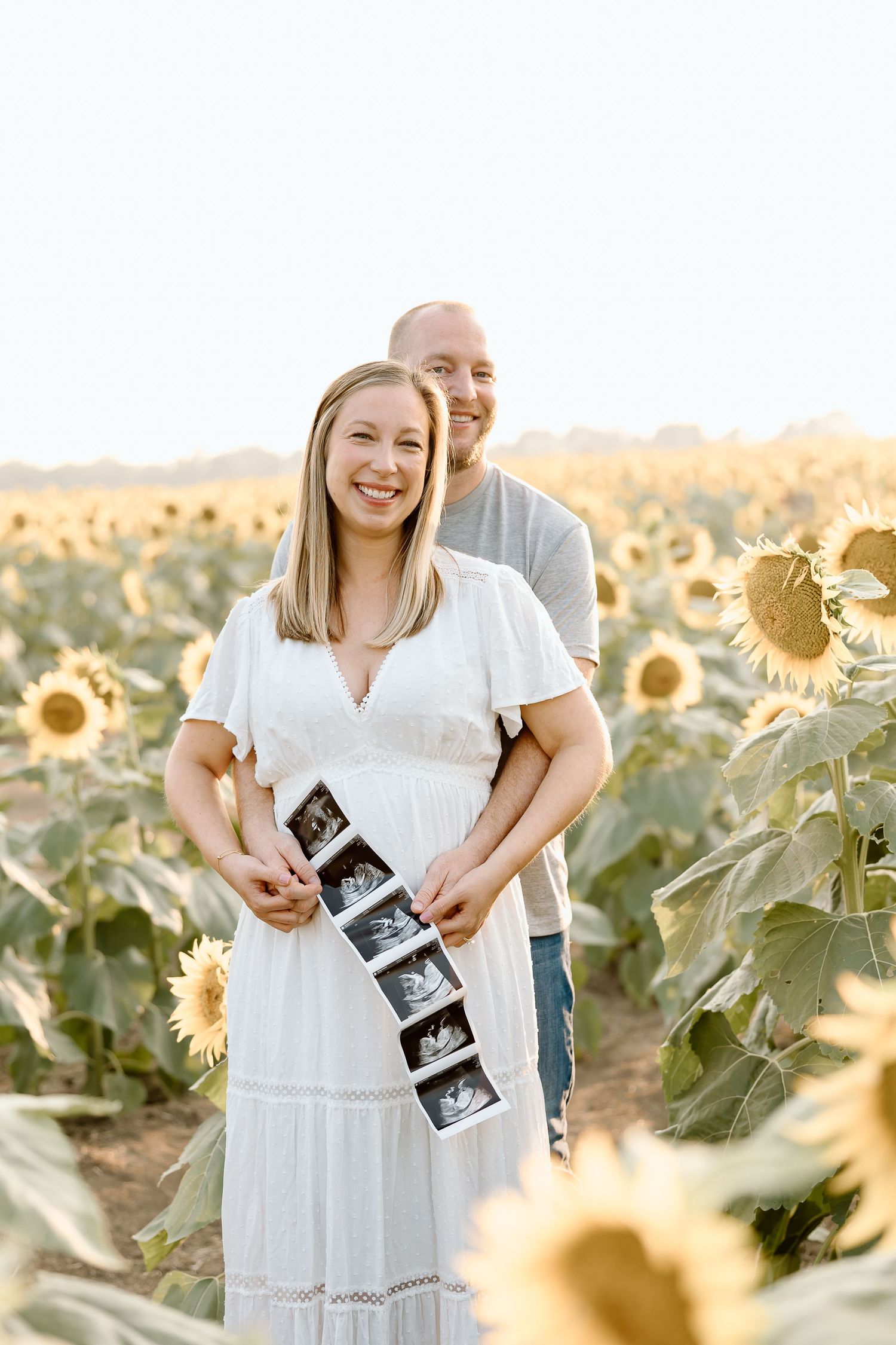 Couple holding ultrasound photos in a sunflower field at sunset, wearing white summer attire.