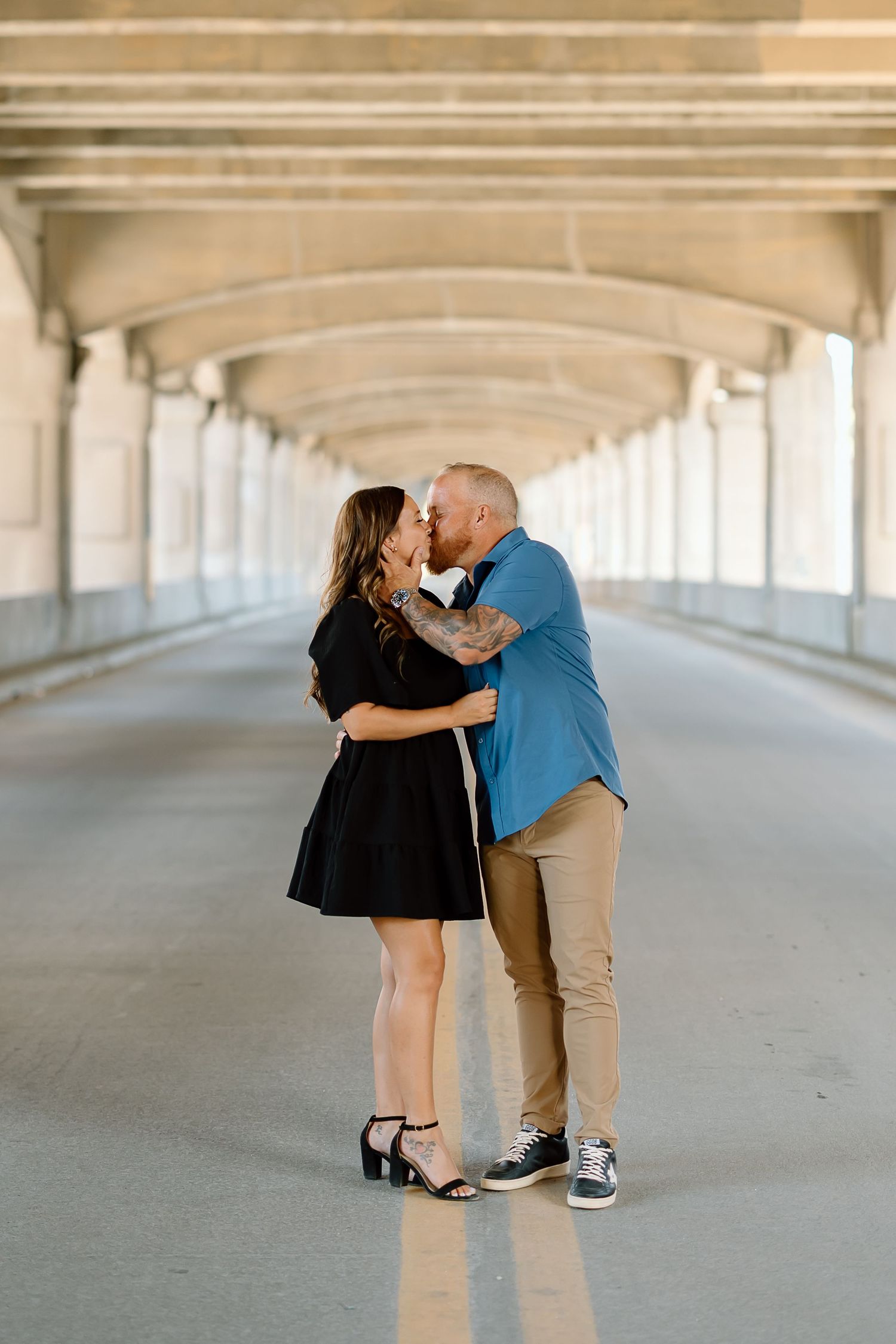 A couple shares a romantic moment under a covered walkway with repeating architectural columns in the background.