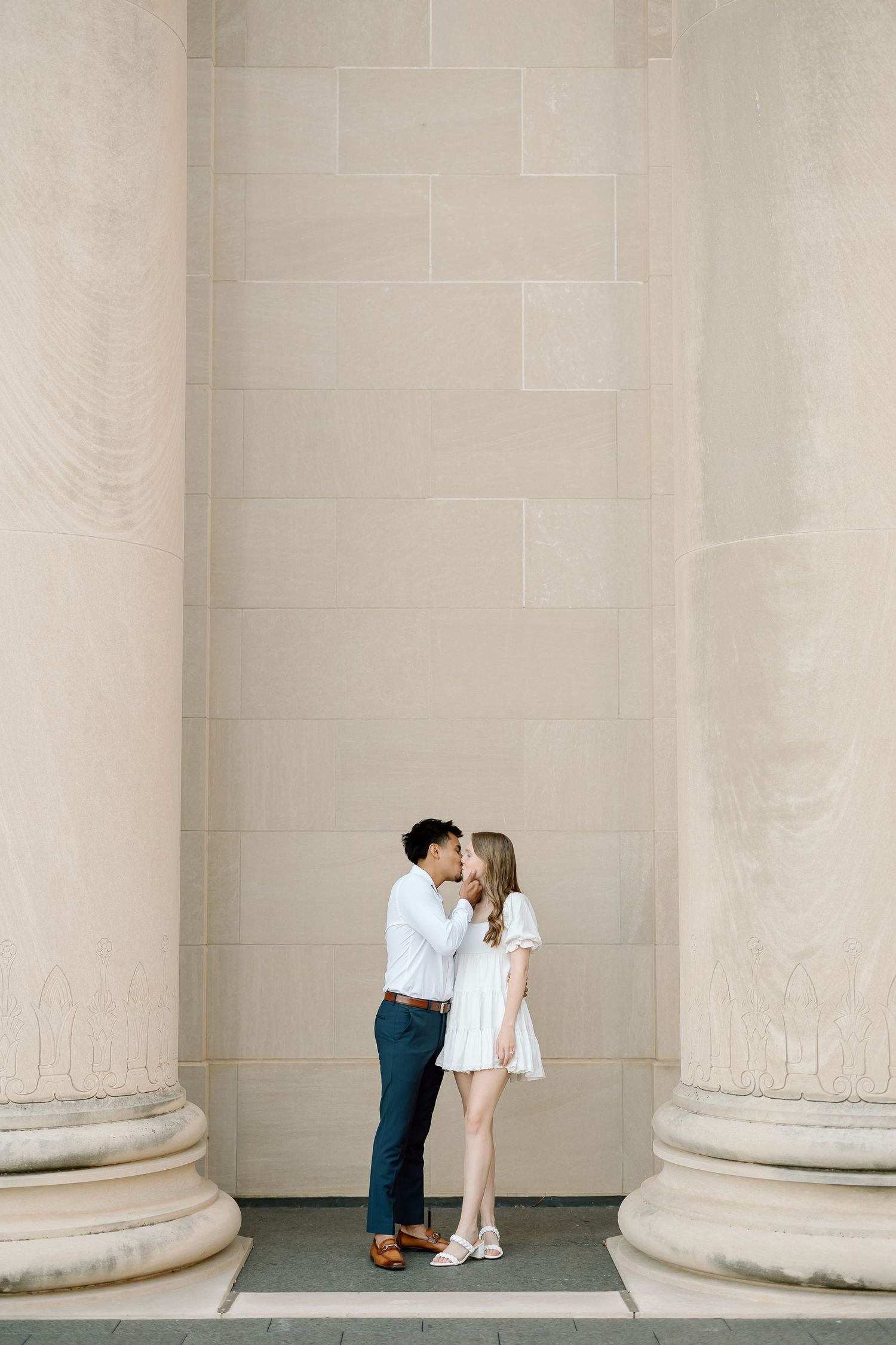 A romantic couple shares a tender moment between classic white stone columns in a minimalist architectural setting.