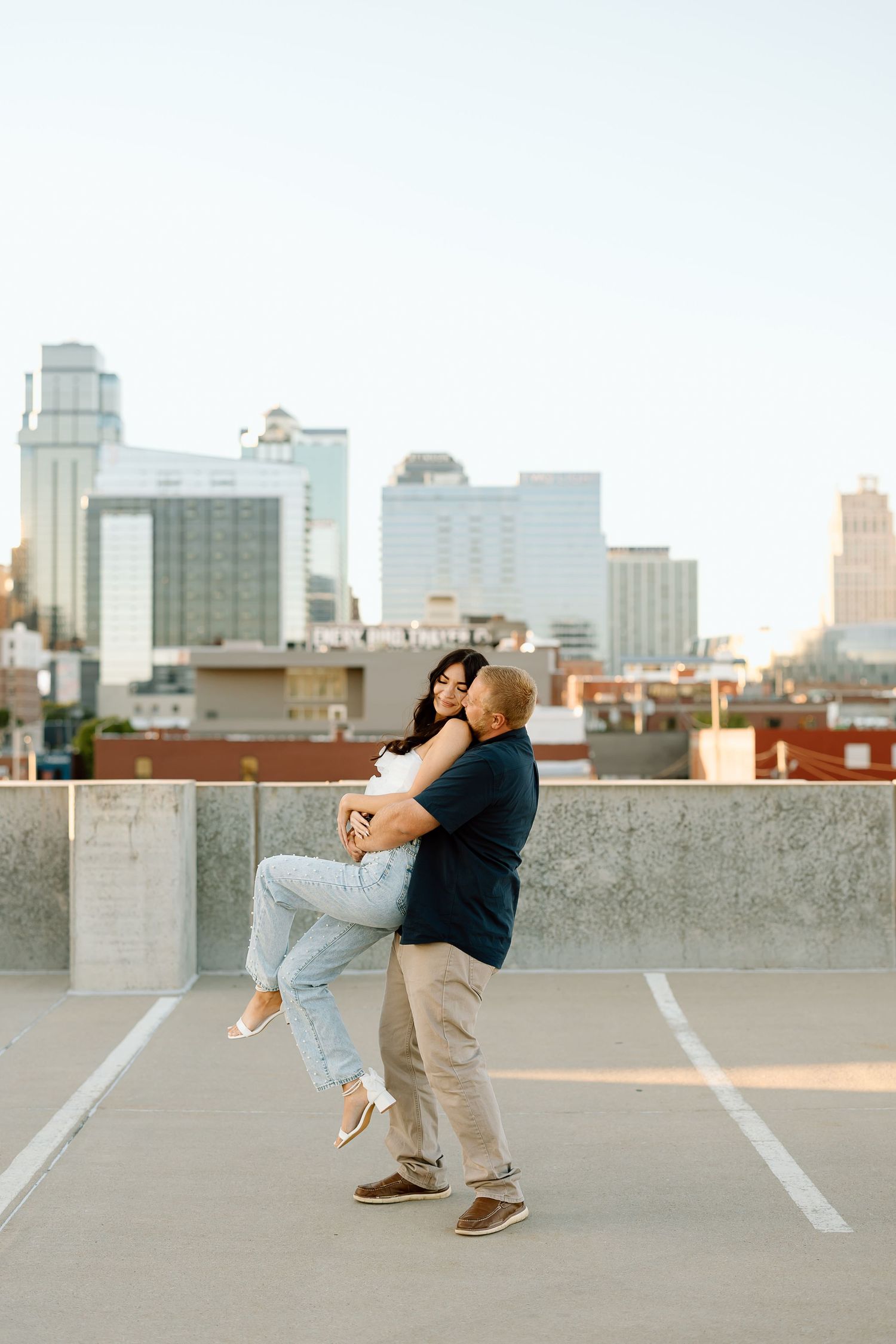 A romantic couple embraces and dances on a rooftop parking lot with a city skyline in the background.