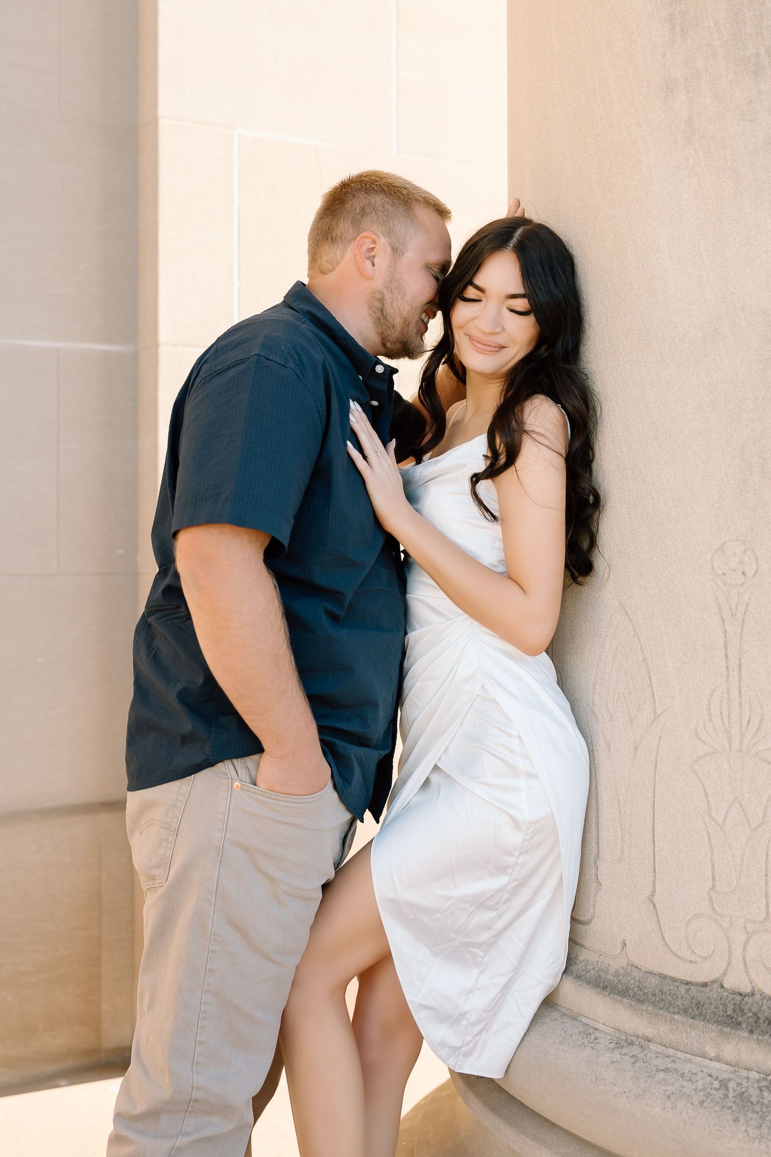 A romantic couple embraces against a light colored wall, with her in a white dress and him in a navy shirt.