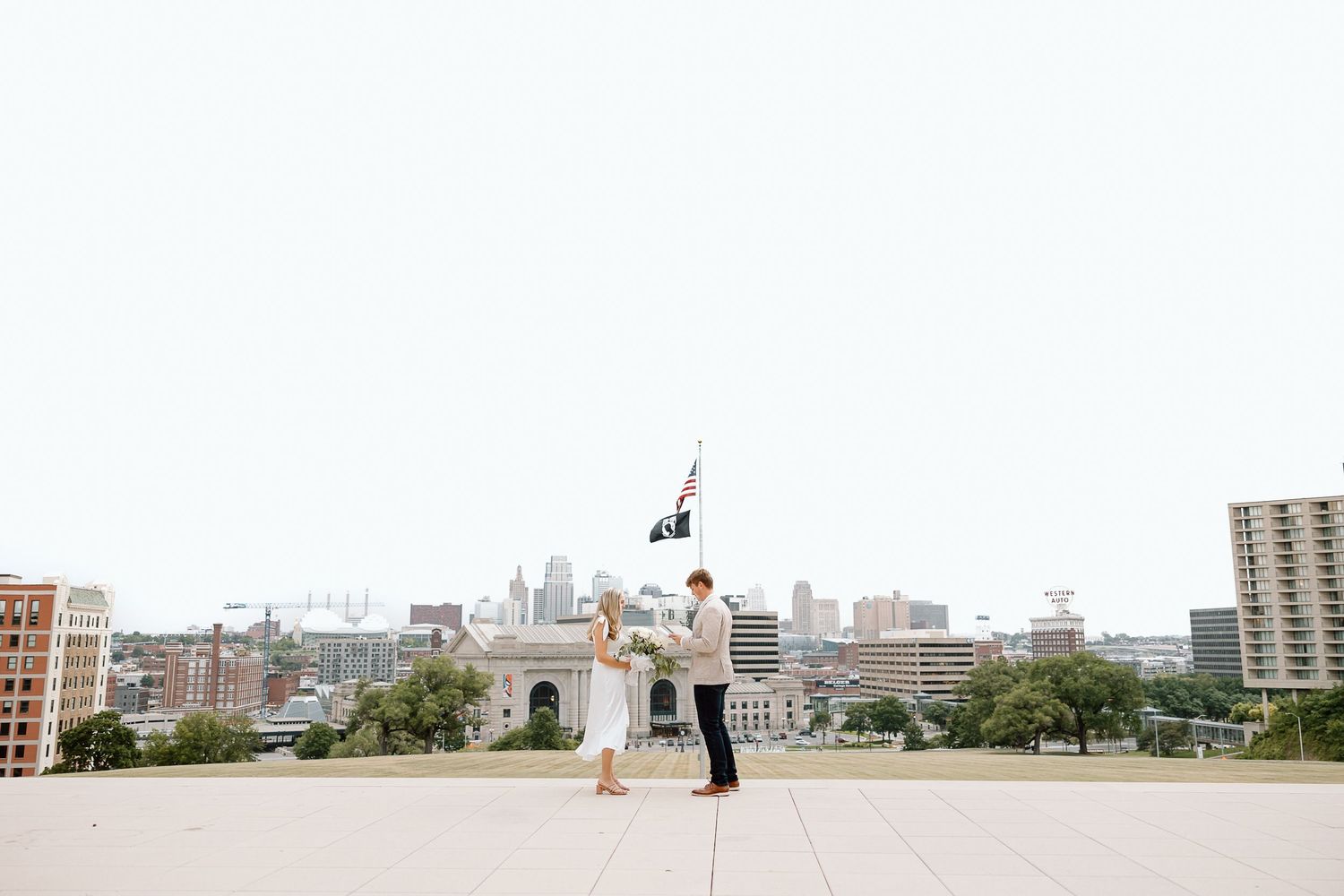 A couple performs an elegant dance lift on a rooftop overlooking a cityscape with a flag pole in the background.