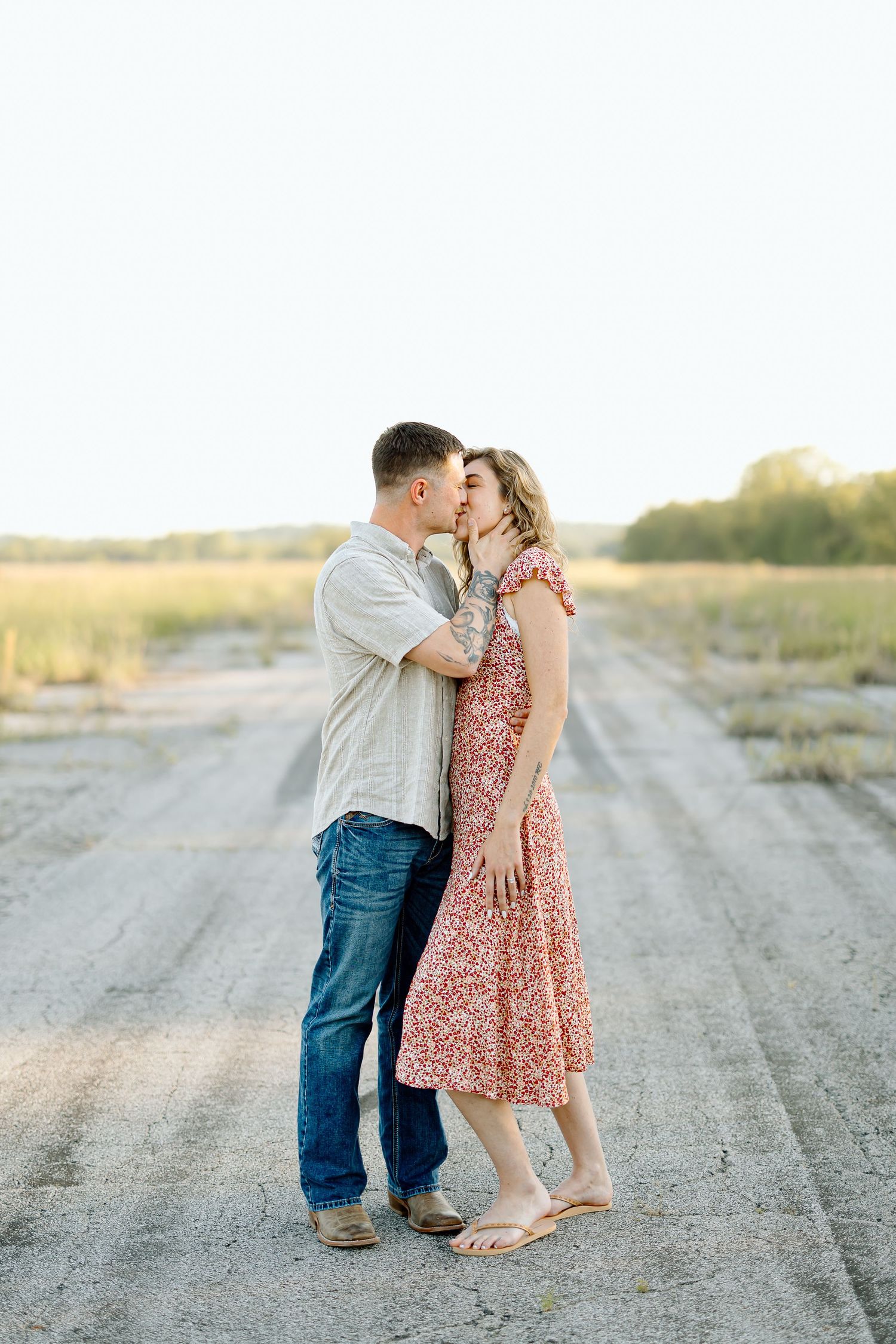 A romantic couple embraces on a dusty rural road at sunset while wearing casual summer attire.