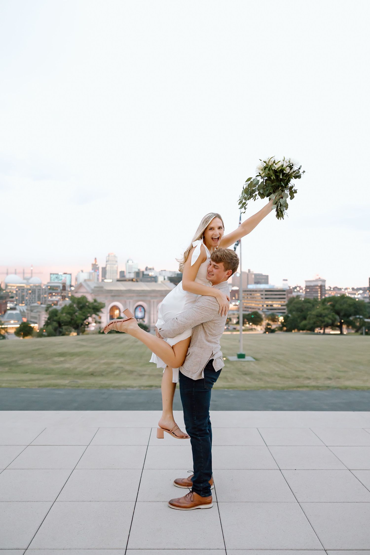 A couple in casual attire dances playfully together in a park with a city skyline visible in the background.