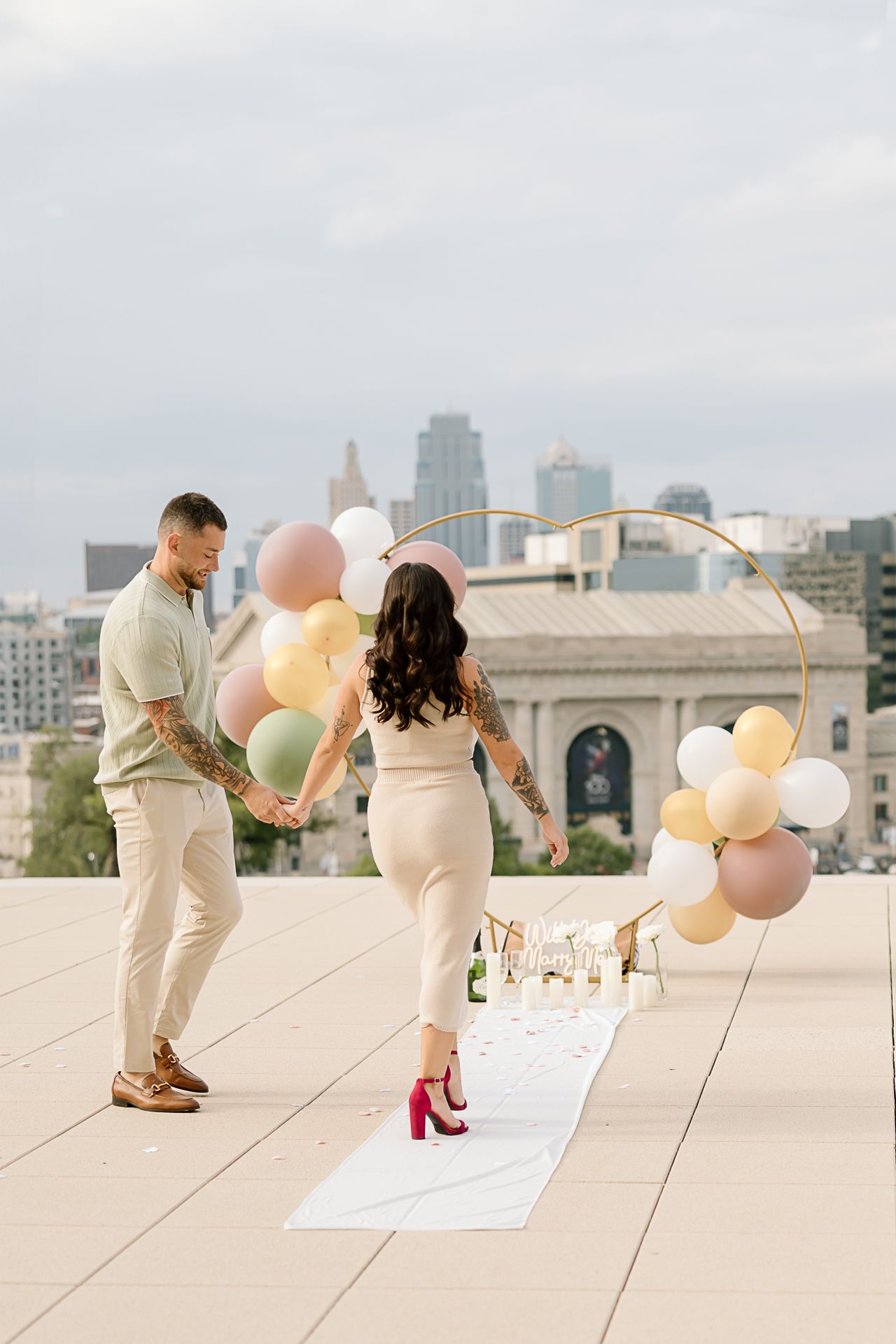 Couple holding pastel balloons on rooftop with city skyline in background during proposal setup.