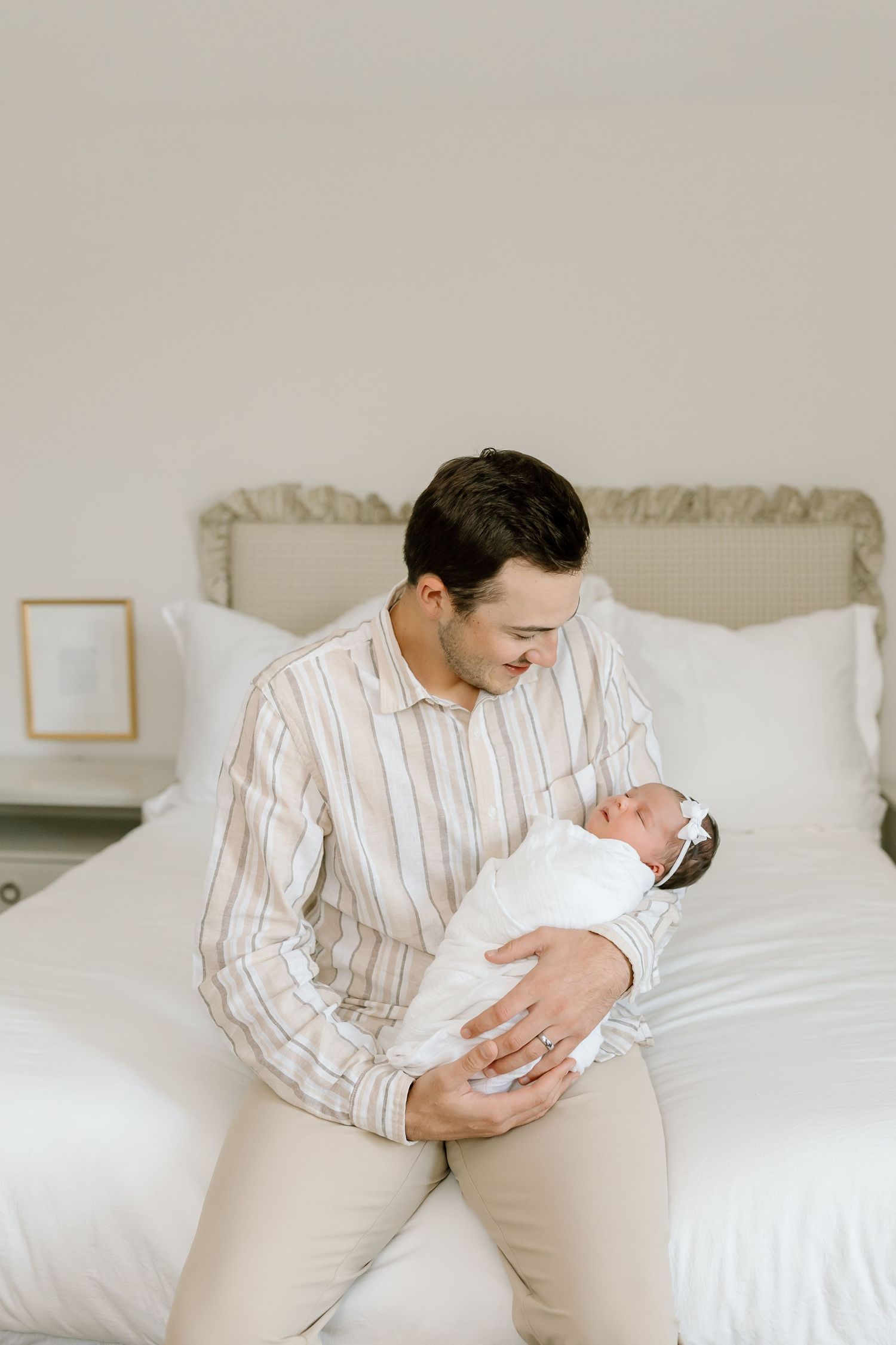 A parent in a striped shirt cradles a newborn baby while sitting on a white bed in a bright, minimalist room.
