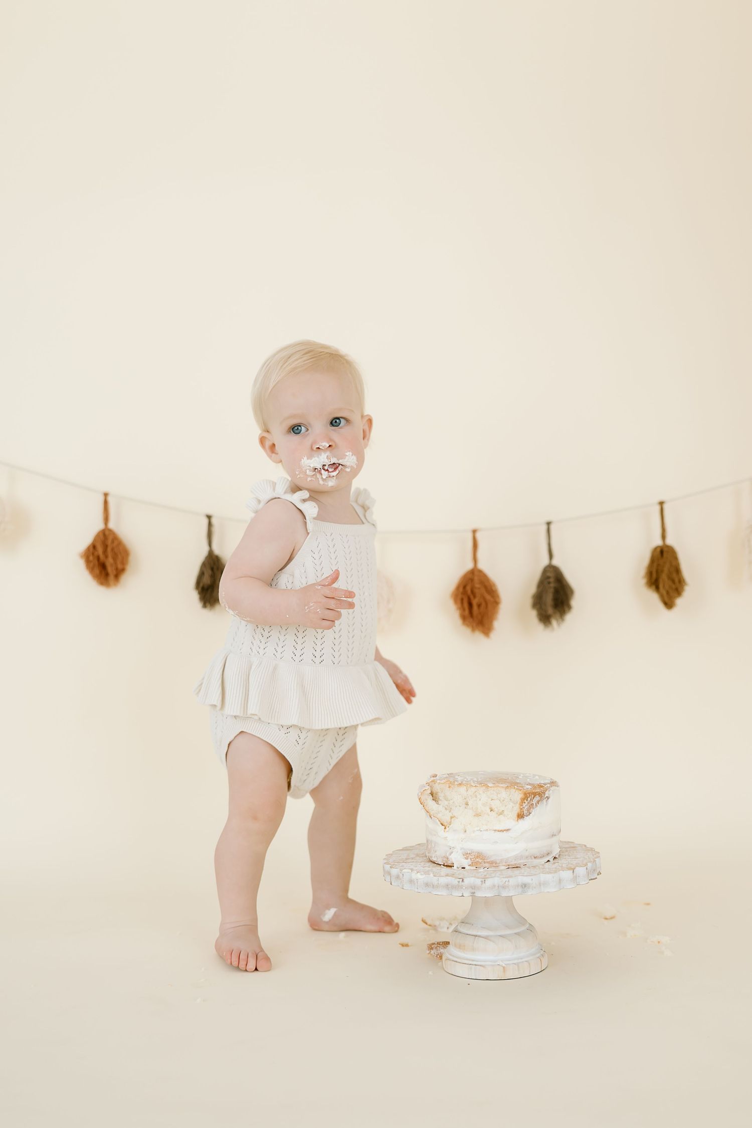 Baby in white outfit plays with cake and feather garland during first birthday cake smash photo session.