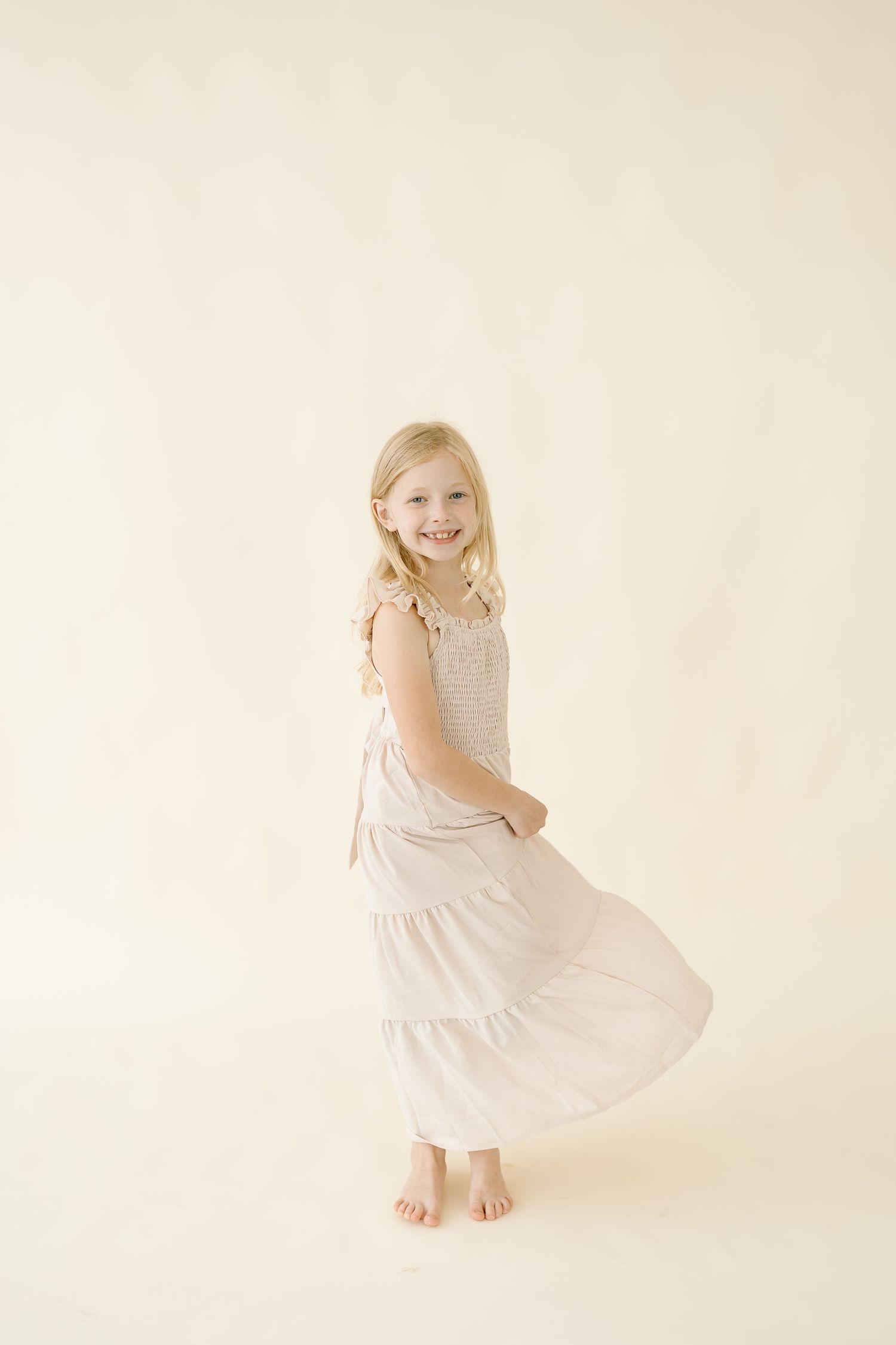 A young child in a white dress twirls and dances joyfully against a light neutral backdrop.