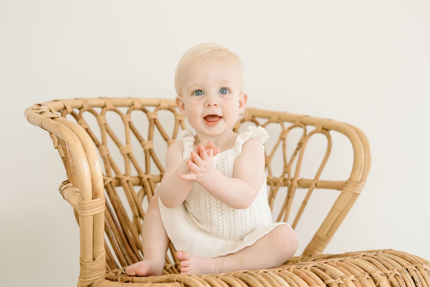 Baby in pink dress sits in woven rattan peacock chair against neutral background for milestone photo session.