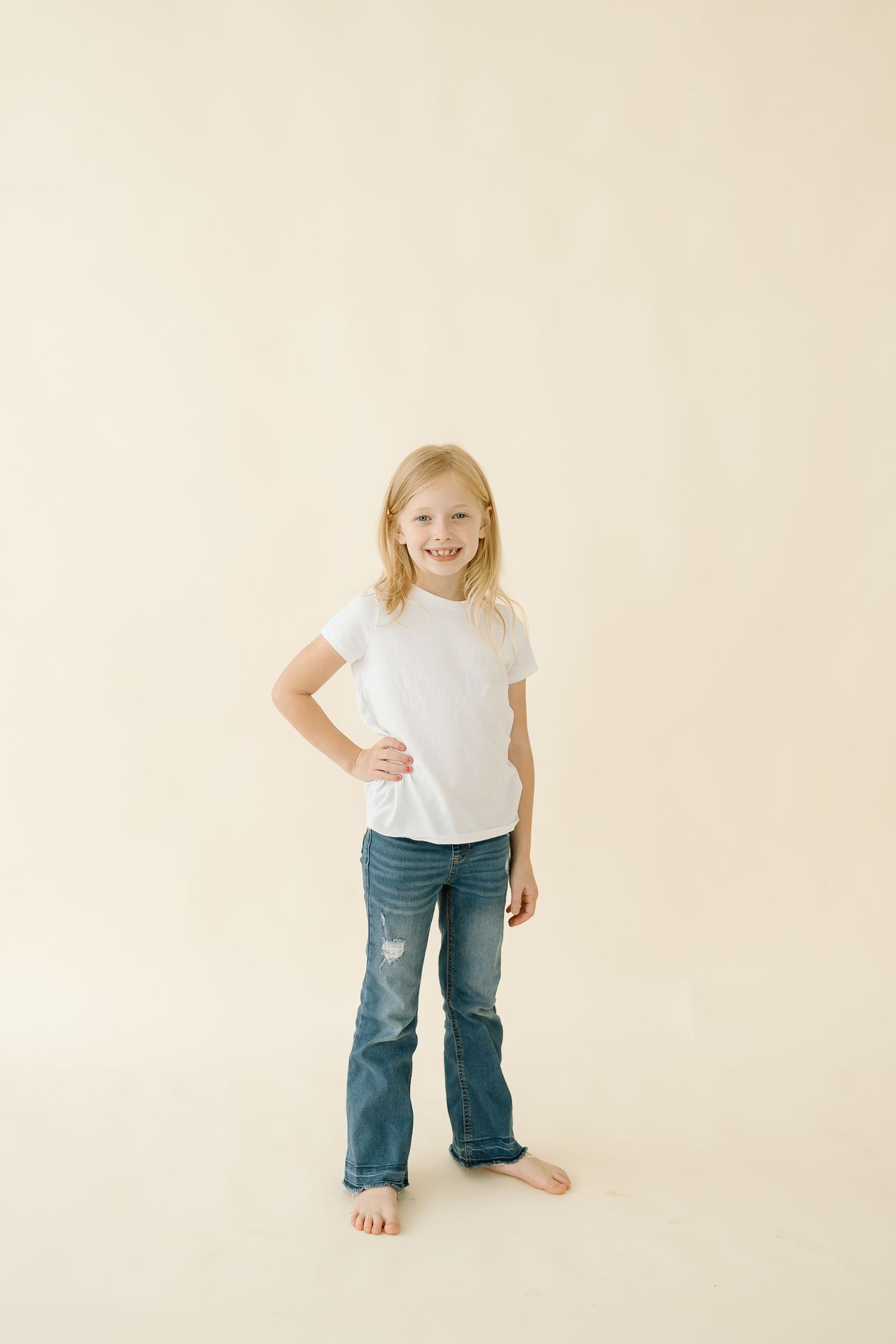 A young child in a white t-shirt and blue jeans poses playfully against a light beige background.