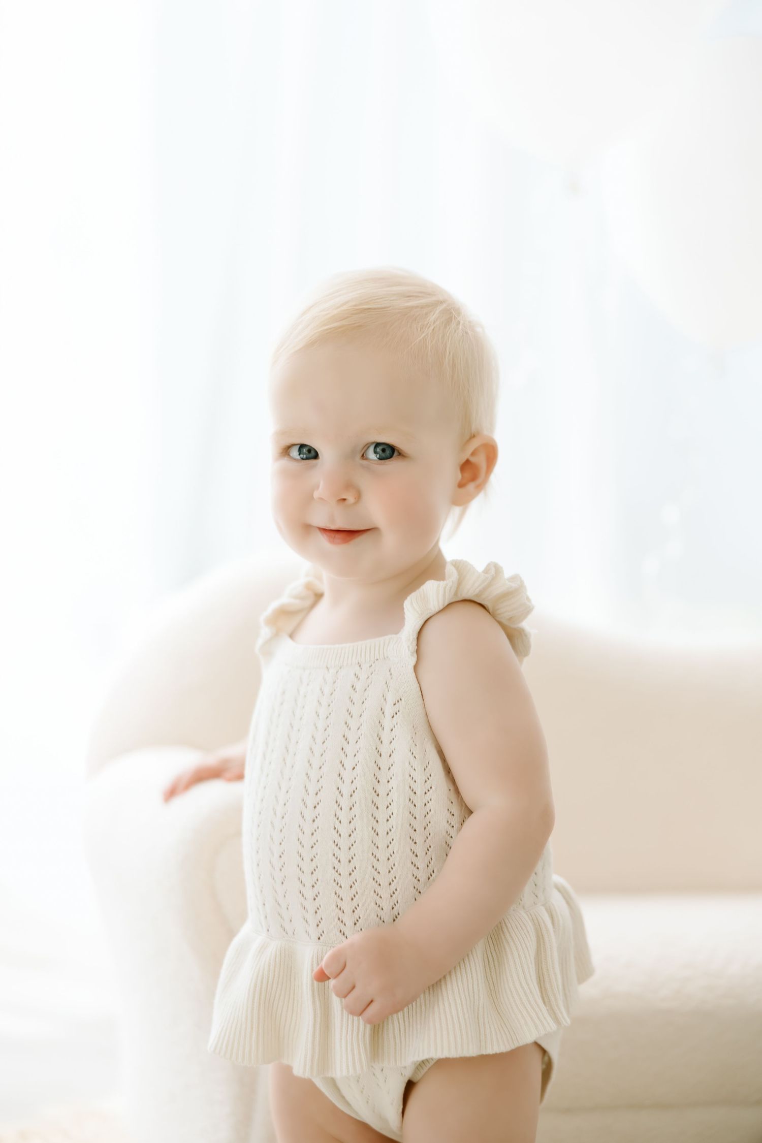 Baby wearing a white knit sleeveless top poses against bright window light in a minimalist setting.
