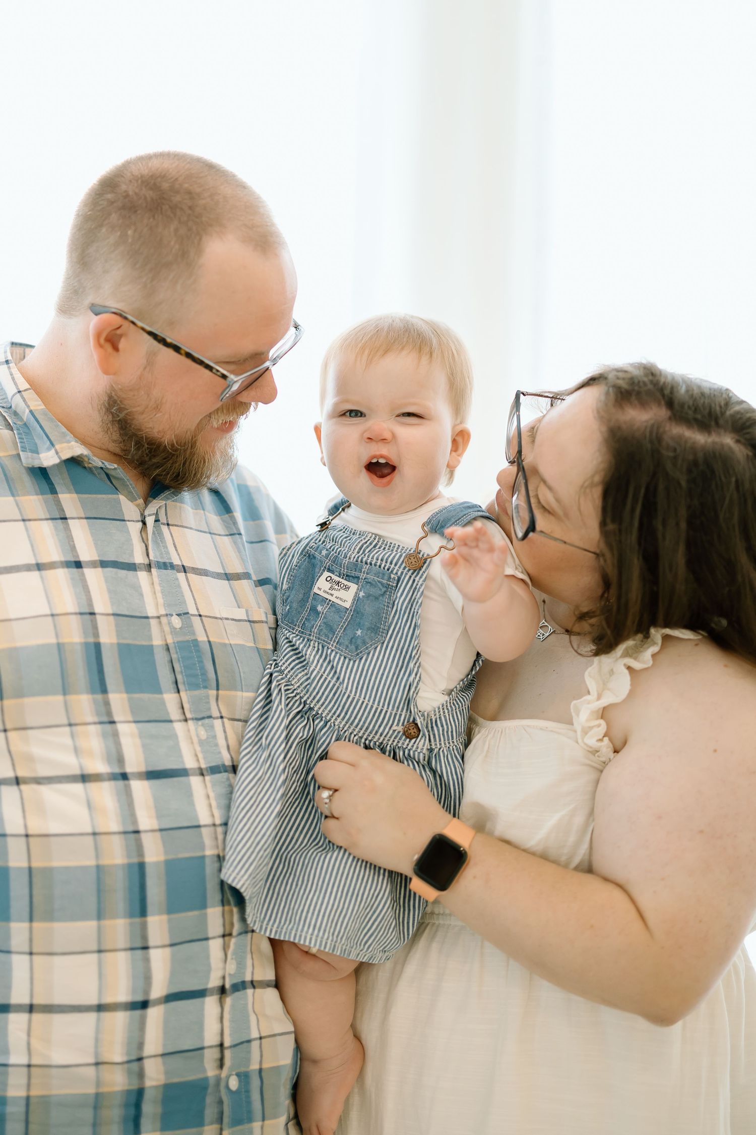 A joyful family moment captured in soft lighting as parents share a laugh with their baby wearing striped overalls.