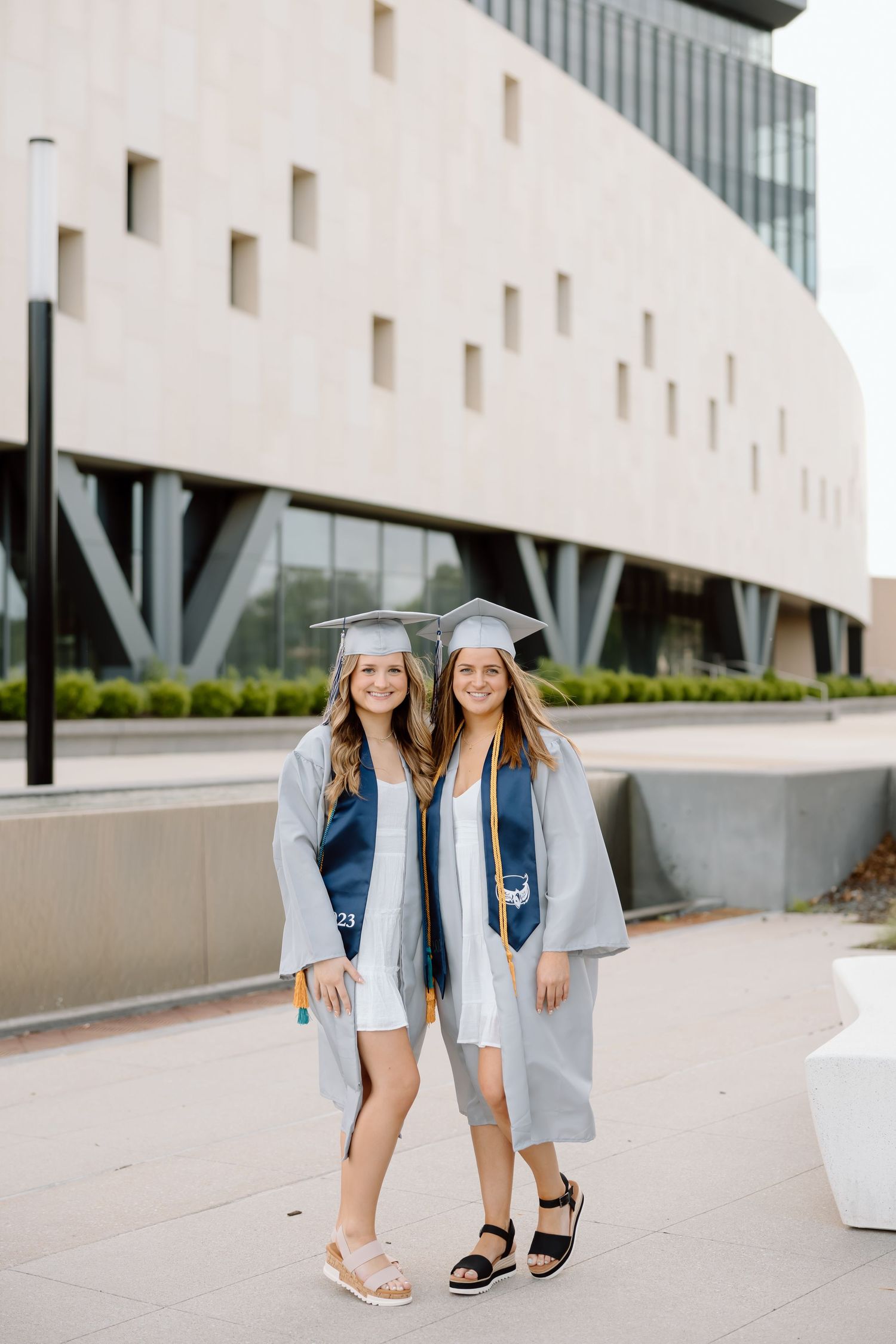 Two graduates in light blue caps and gowns pose together on a modern campus walkway.