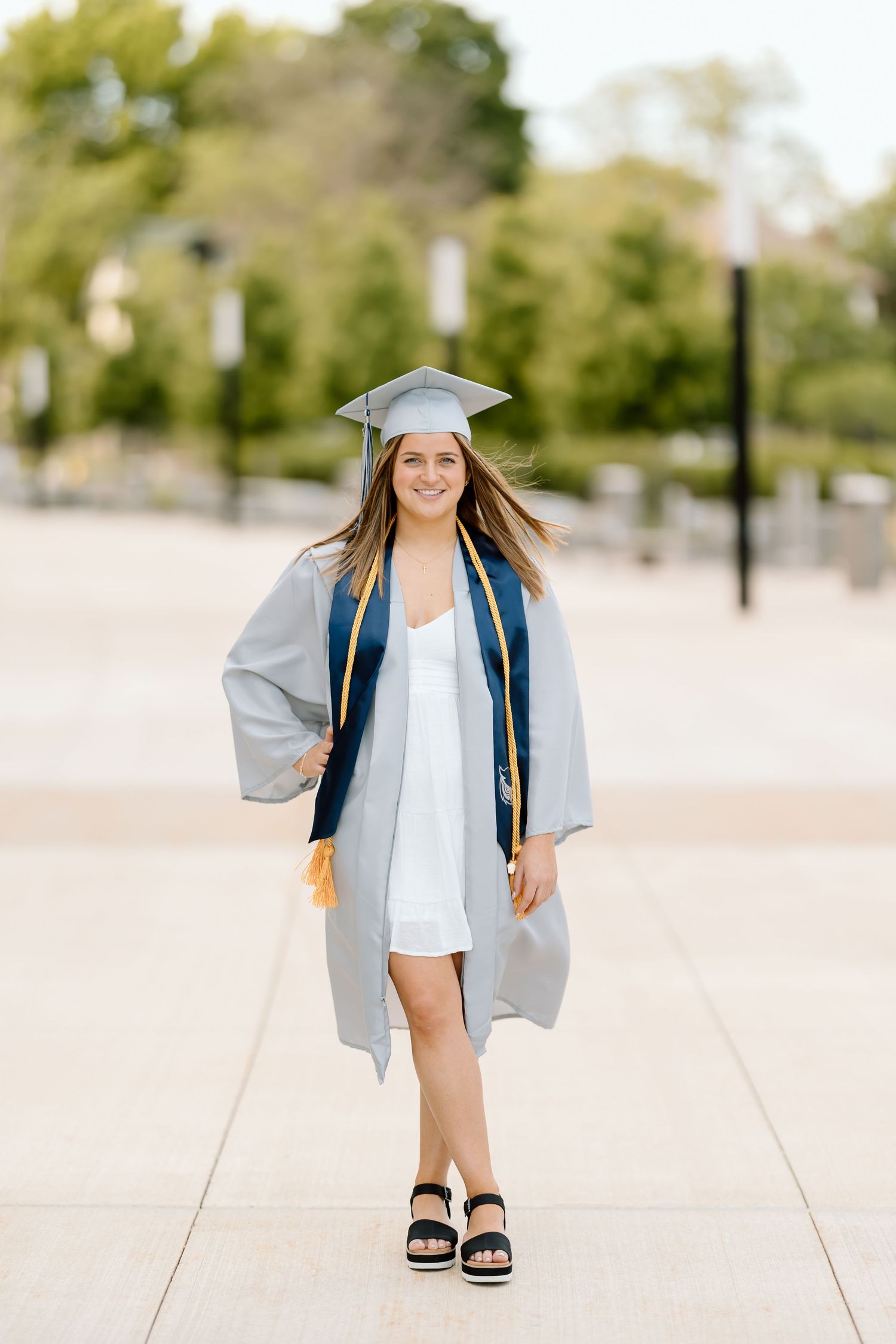 Graduate in blue cap and gown poses outdoors on sunny day with trees in background.