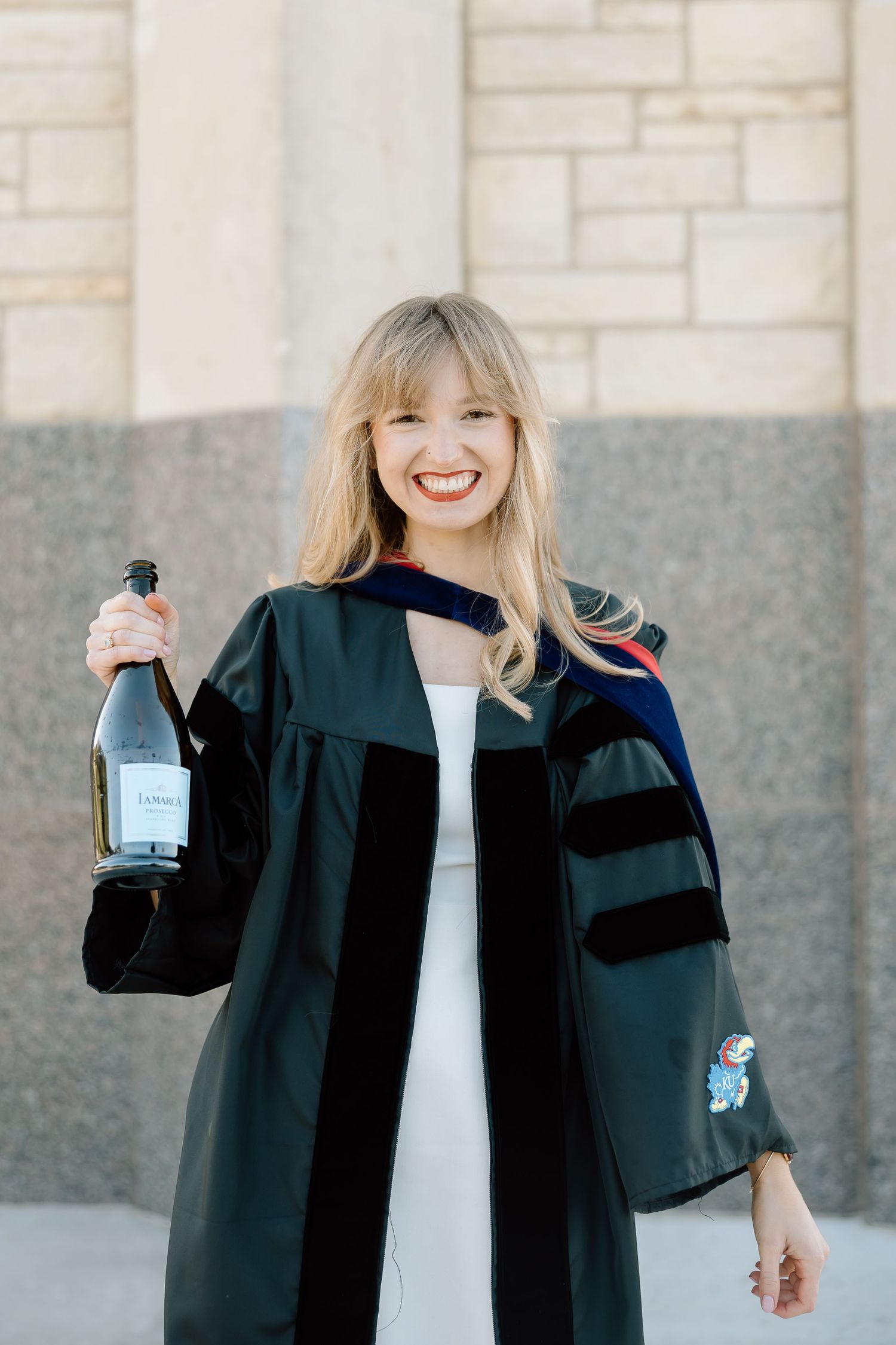 Graduate wearing black academic regalia holds up diploma while celebrating outdoors against stone wall background.