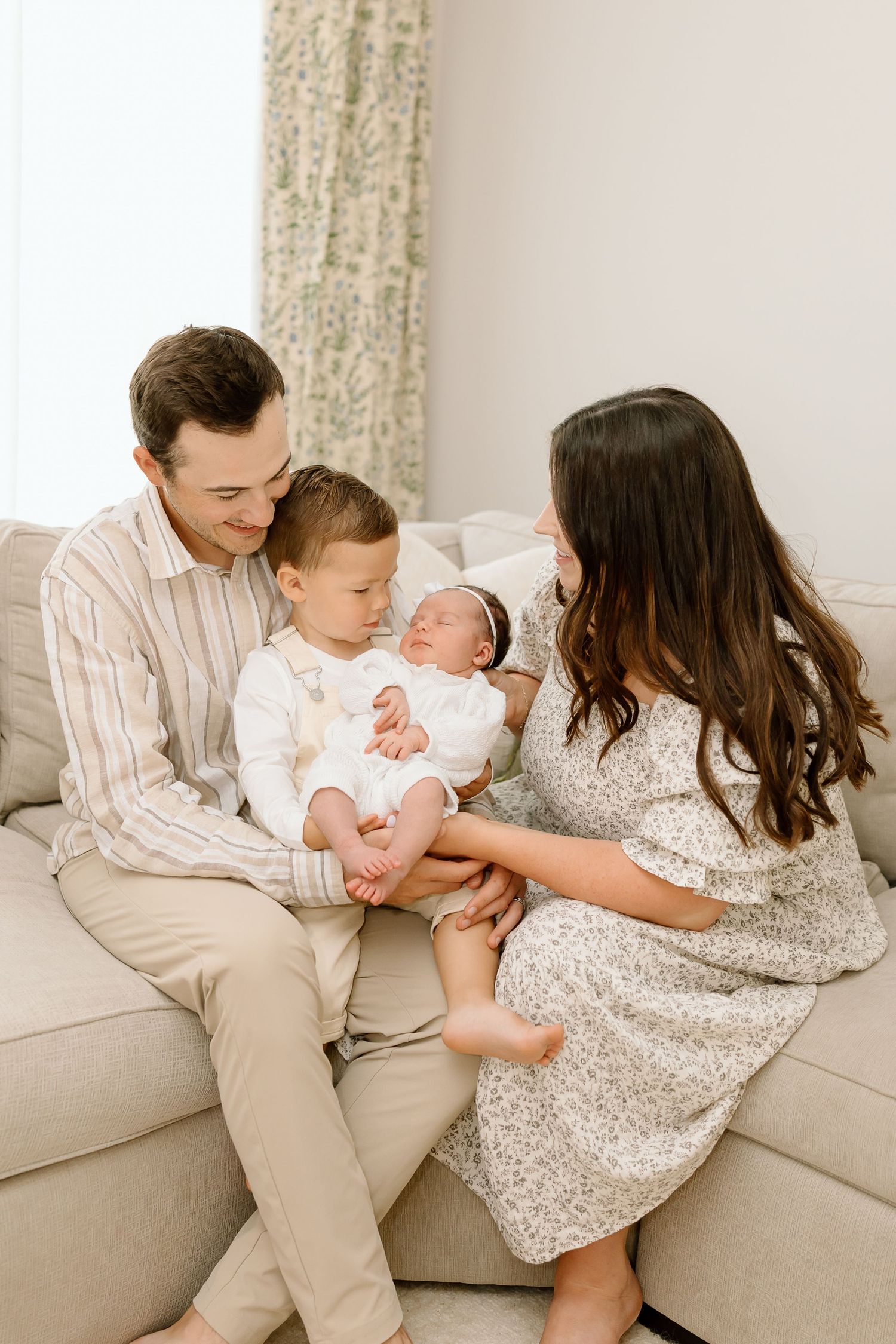 A family of four sits together on a beige couch sharing tender moments with their newborn in a series of photos.