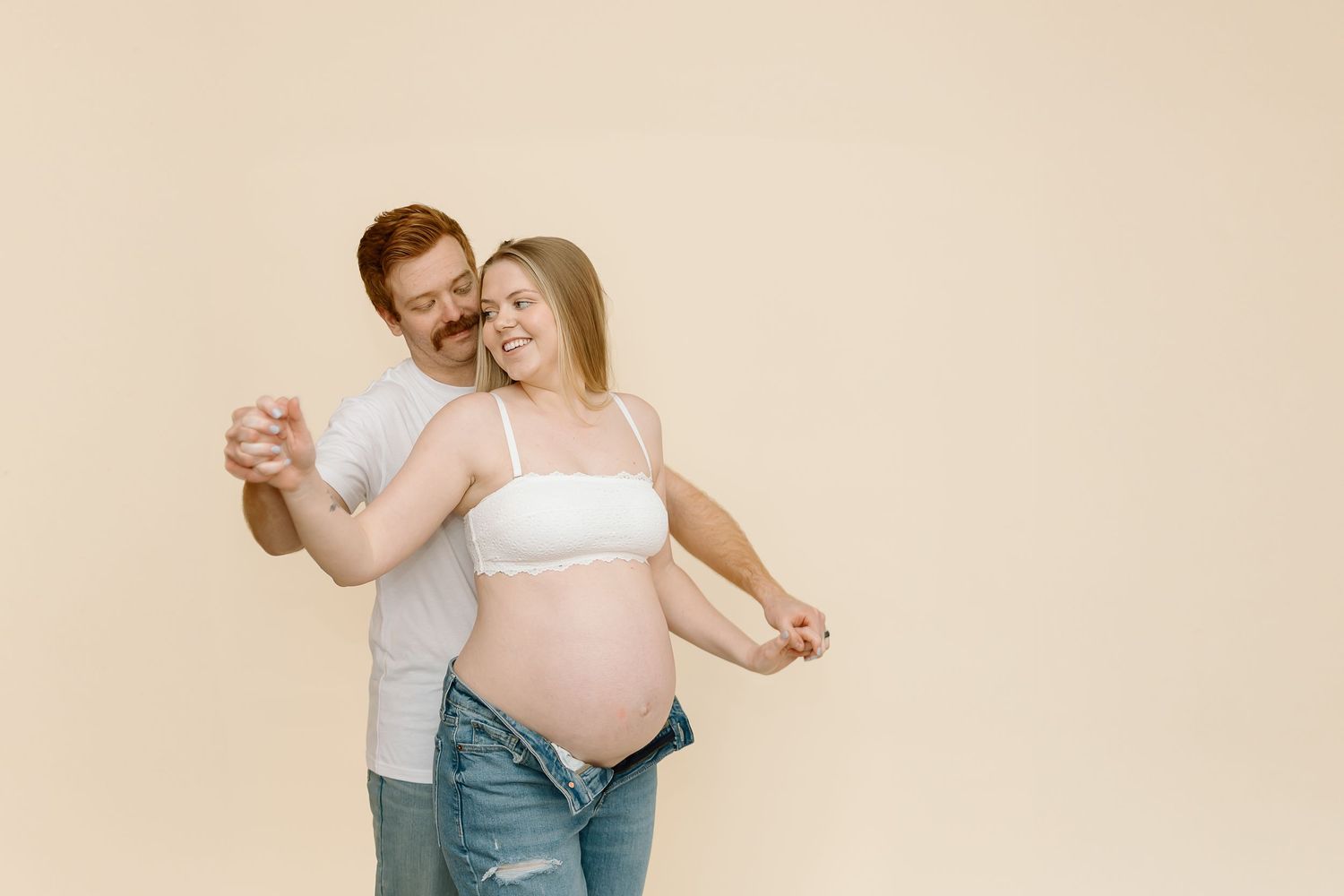 Tender maternity dance pose against beige backdrop captures couple's joy during pregnancy.