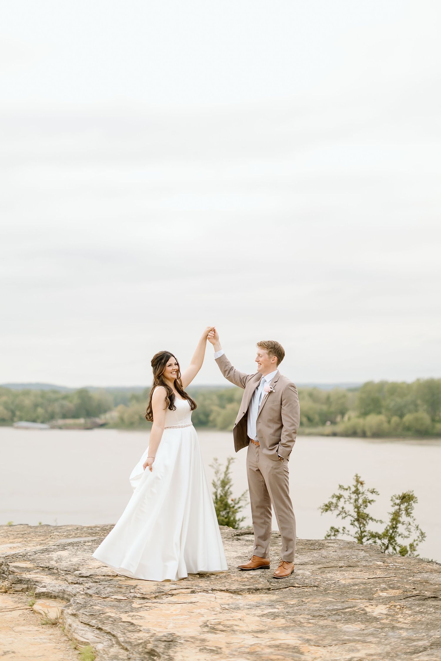 Joyful wedding couple celebrates on rocky overlook with lake view.