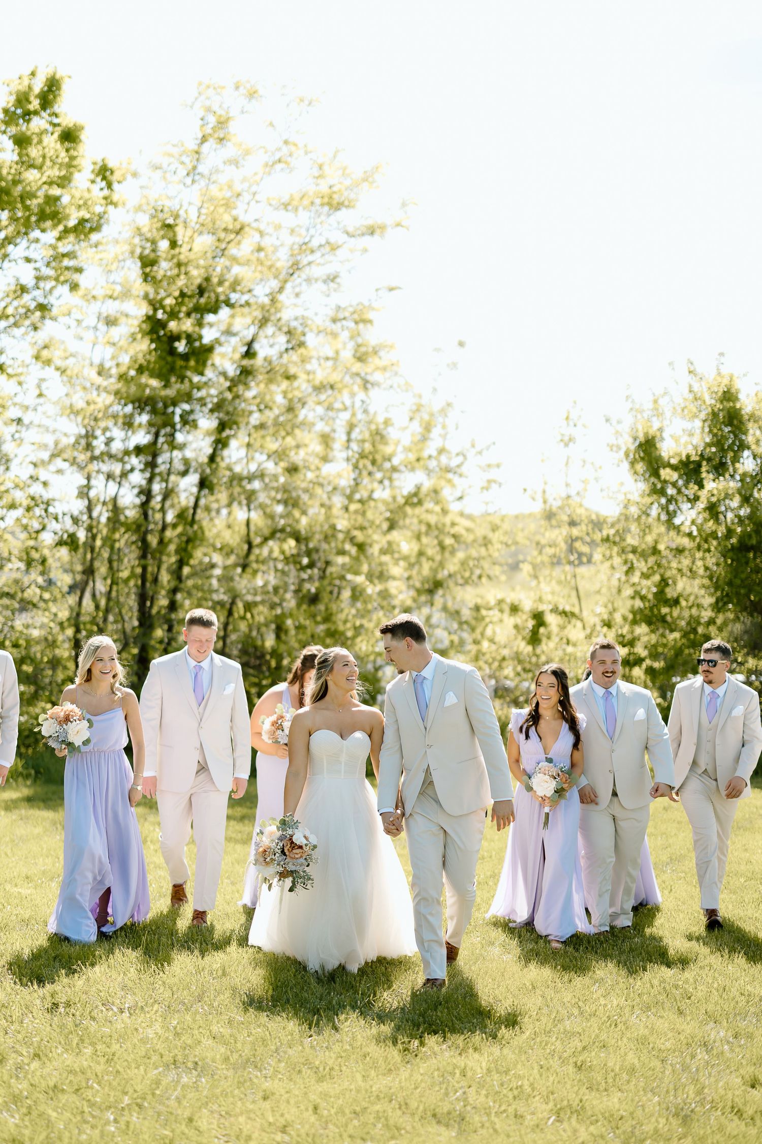 Wedding party walks together on grassy hill with bridesmaids in blue dresses and groomsmen in beige suits.