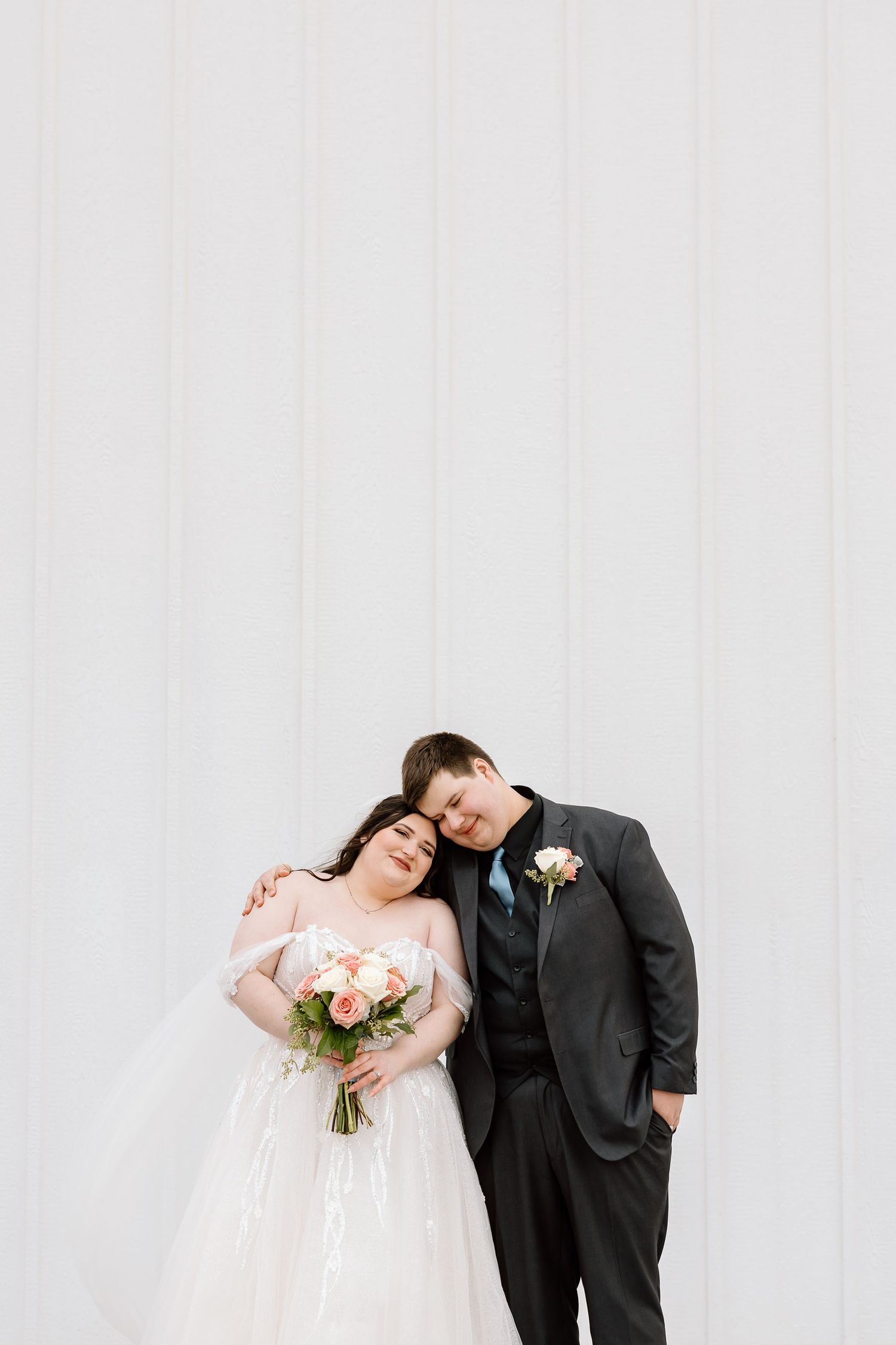 A romantic wedding portrait against a white wall with the couple sharing a tender moment and holding a peach bouquet.