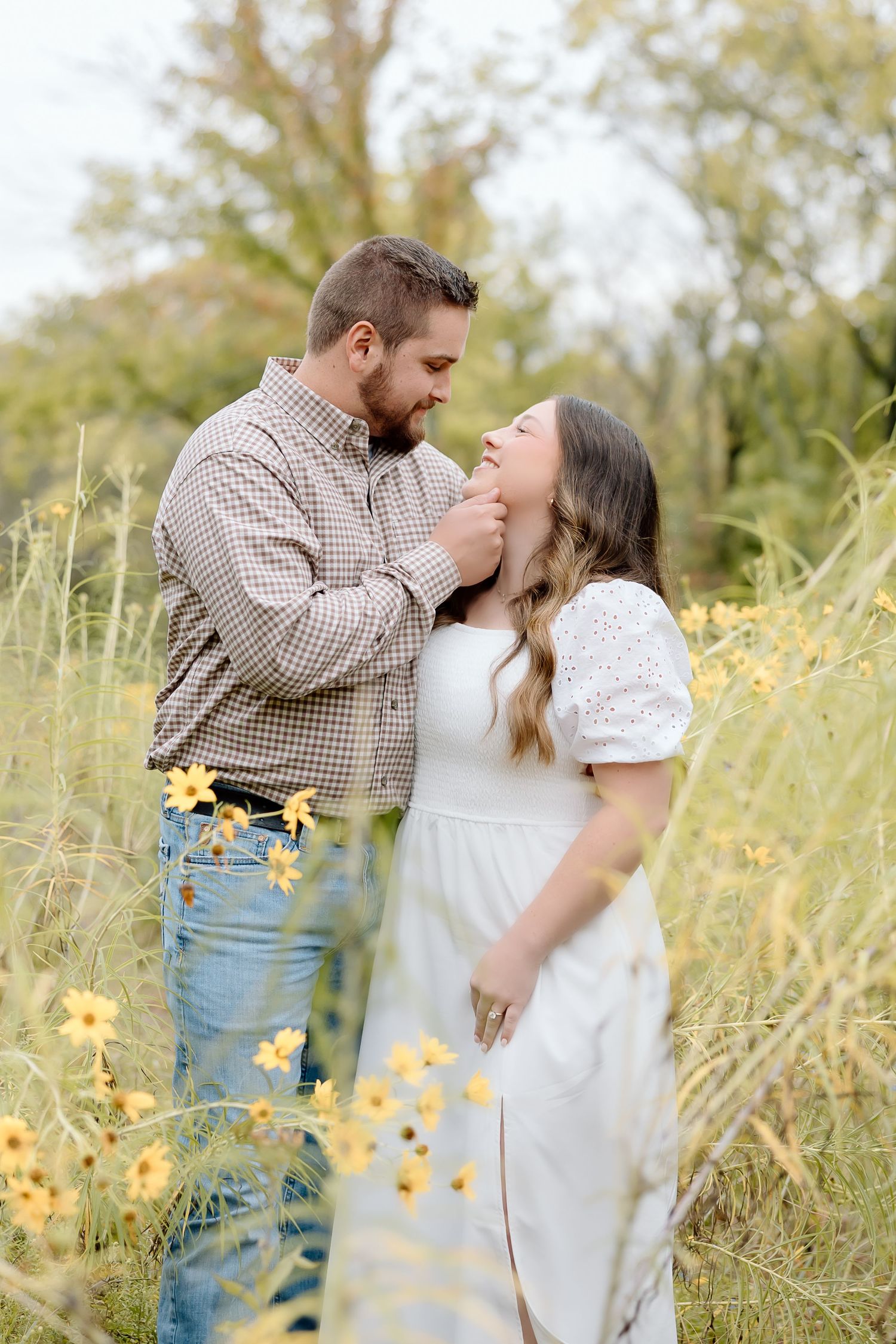 A romantic couple embraces in a meadow filled with yellow wildflowers on a sunny afternoon.