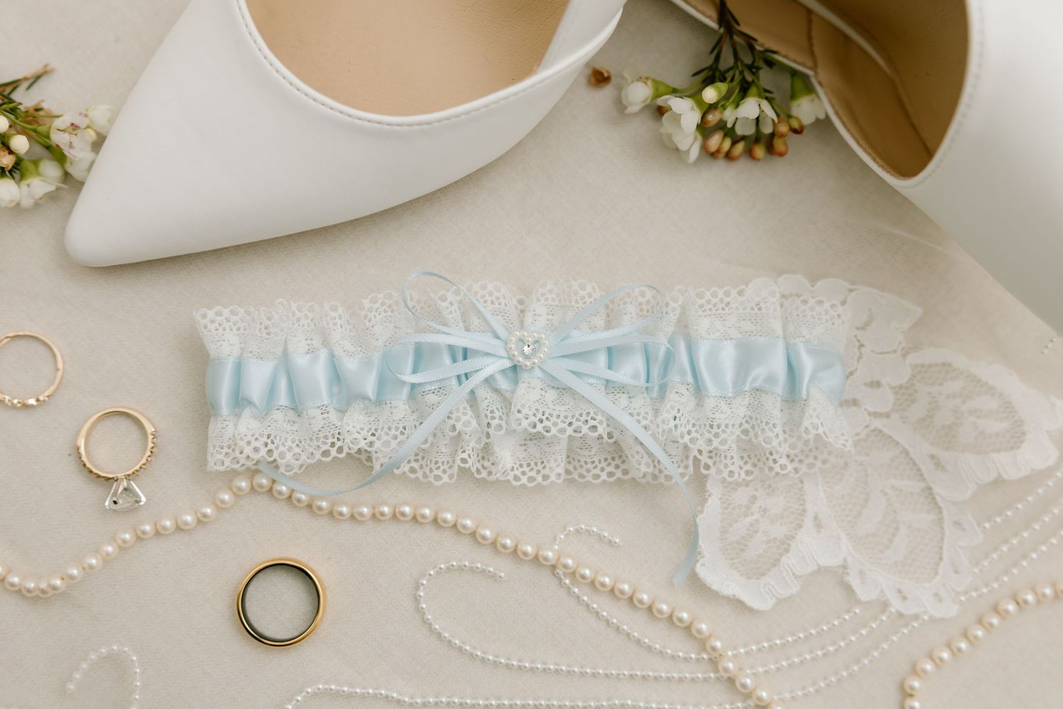 Blue and white lace wedding garter with gold rings and pearl necklace on neutral fabric.