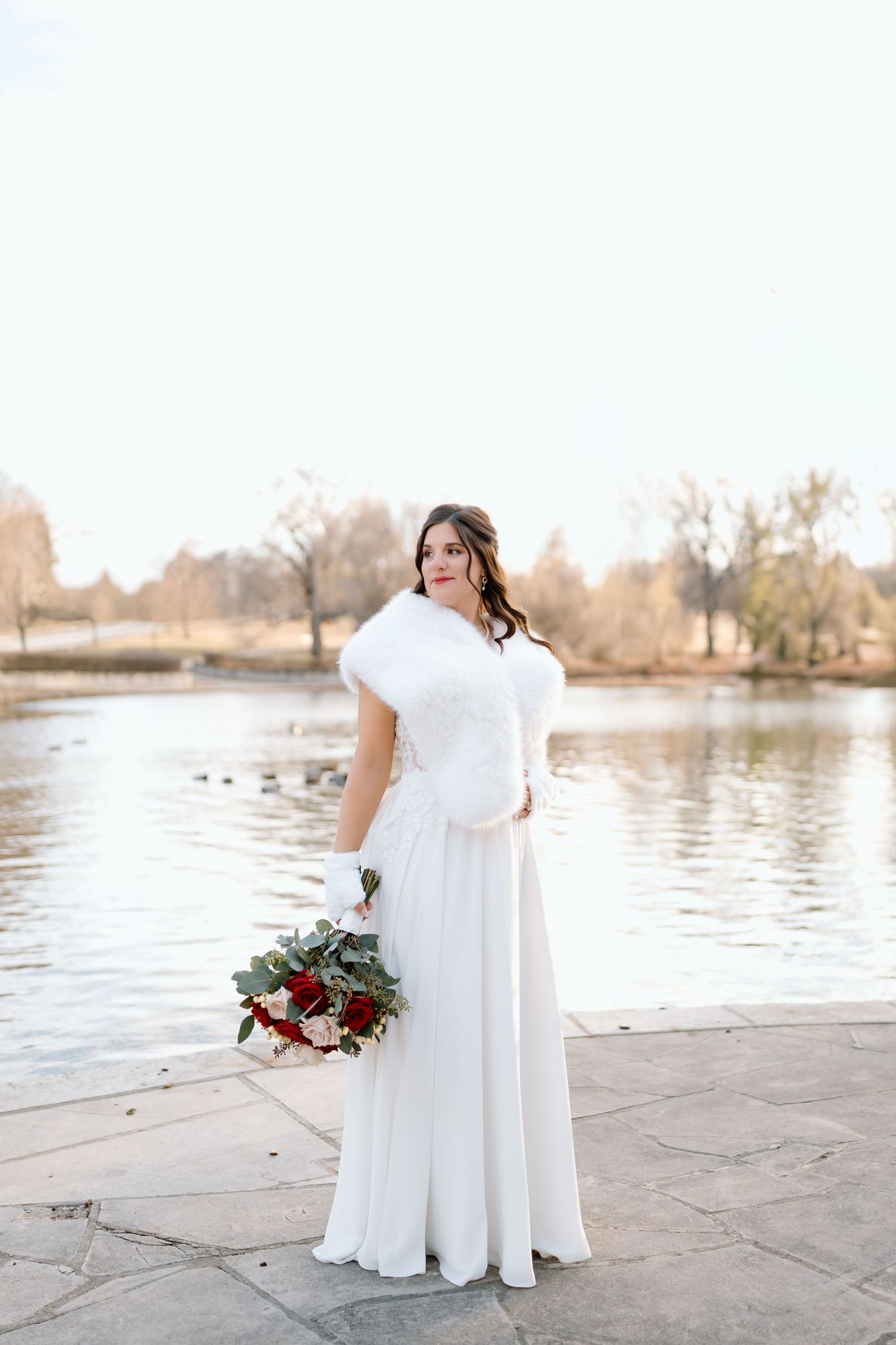 A person in a flowing white dress stands by a serene lake holding a burgundy and green floral bouquet.