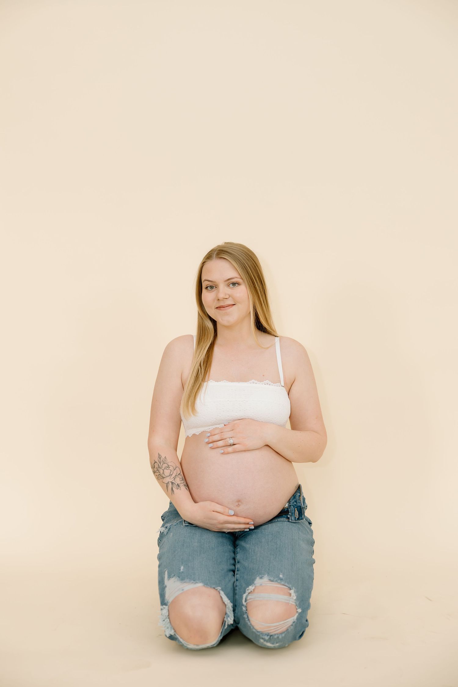 An expecting mother in ripped jeans kneels while cradling her baby bump against a neutral backdrop.