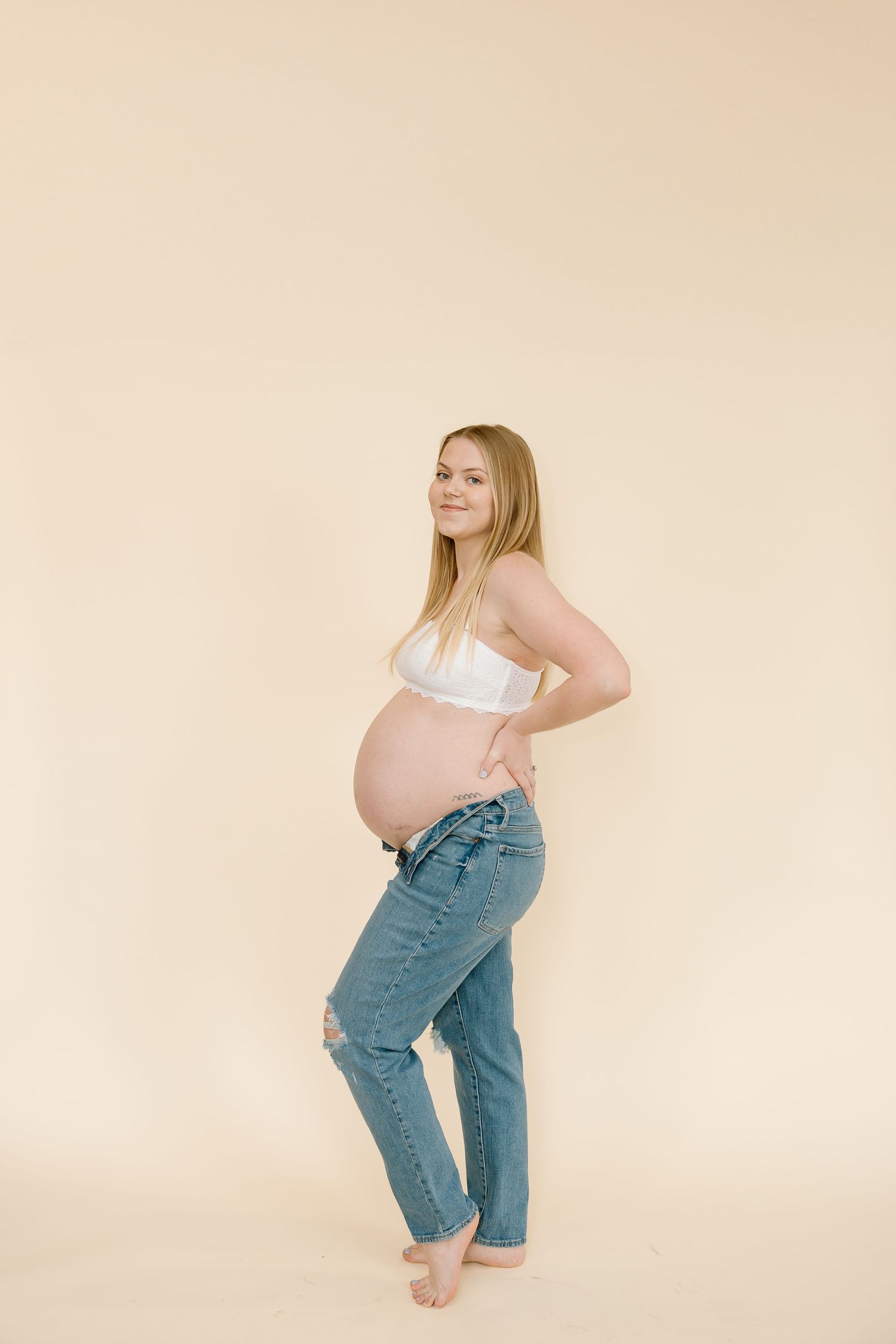 Expectant mother in white top and jeans poses confidently against beige background.