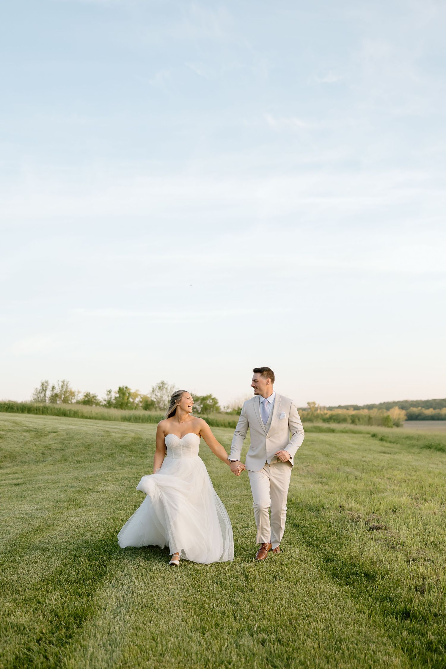 Bride and groom walk together on a grassy hill during golden hour photography session.