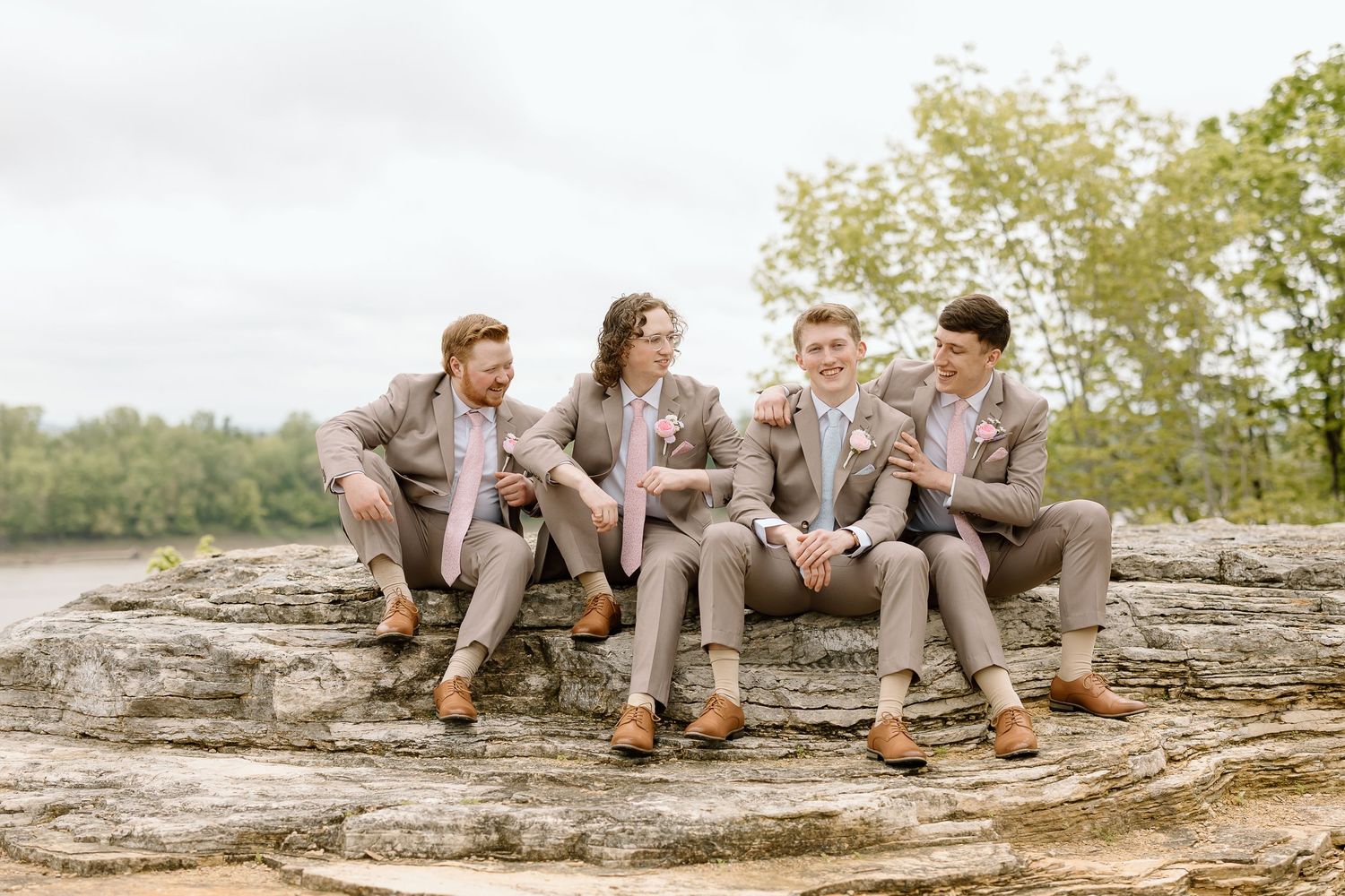 Wedding party in beige attire sits casually on rocky outcrop surrounded by nature.