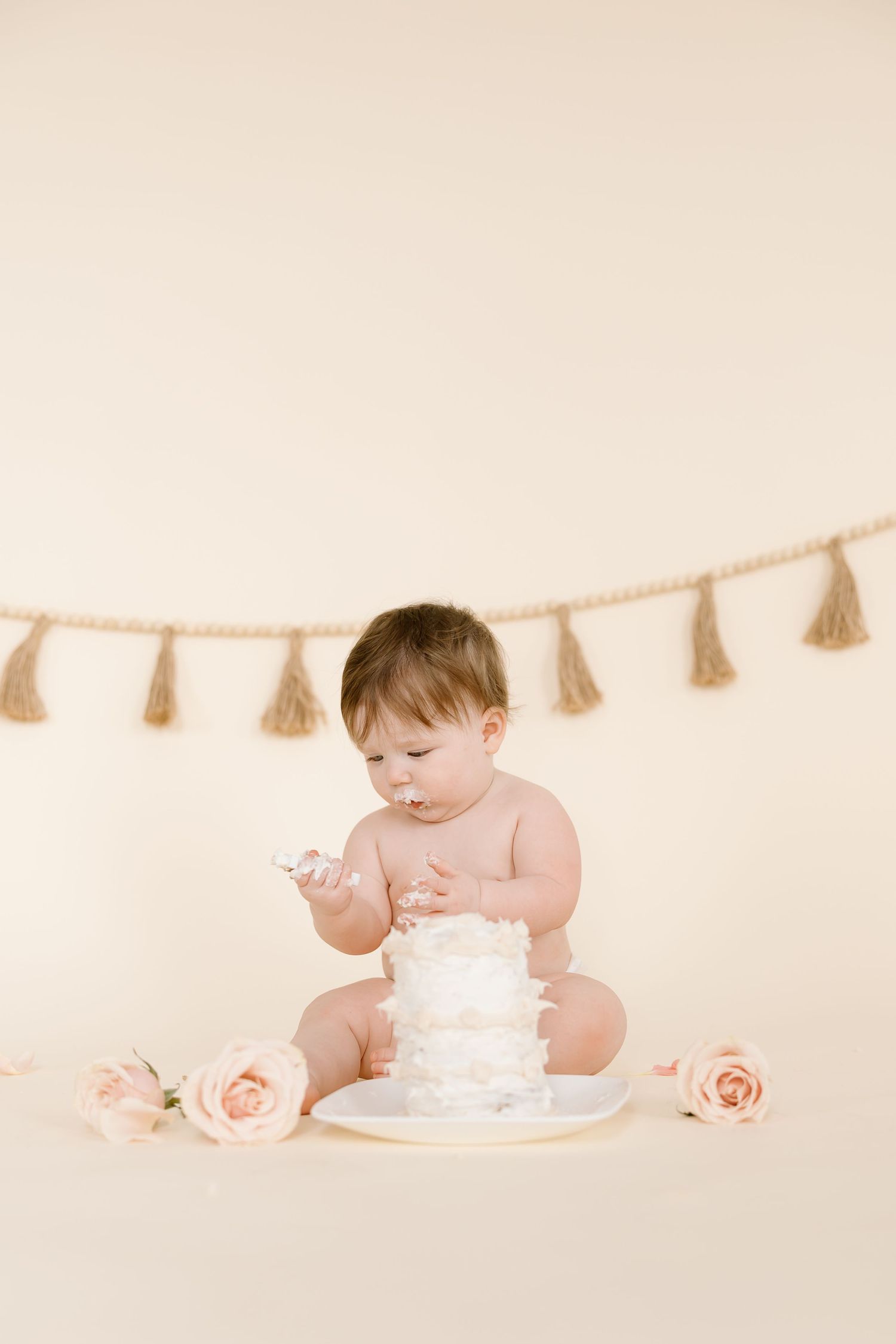 A baby in a diaper sits with a small white cake and pink roses against a beige backdrop with tassel garland.